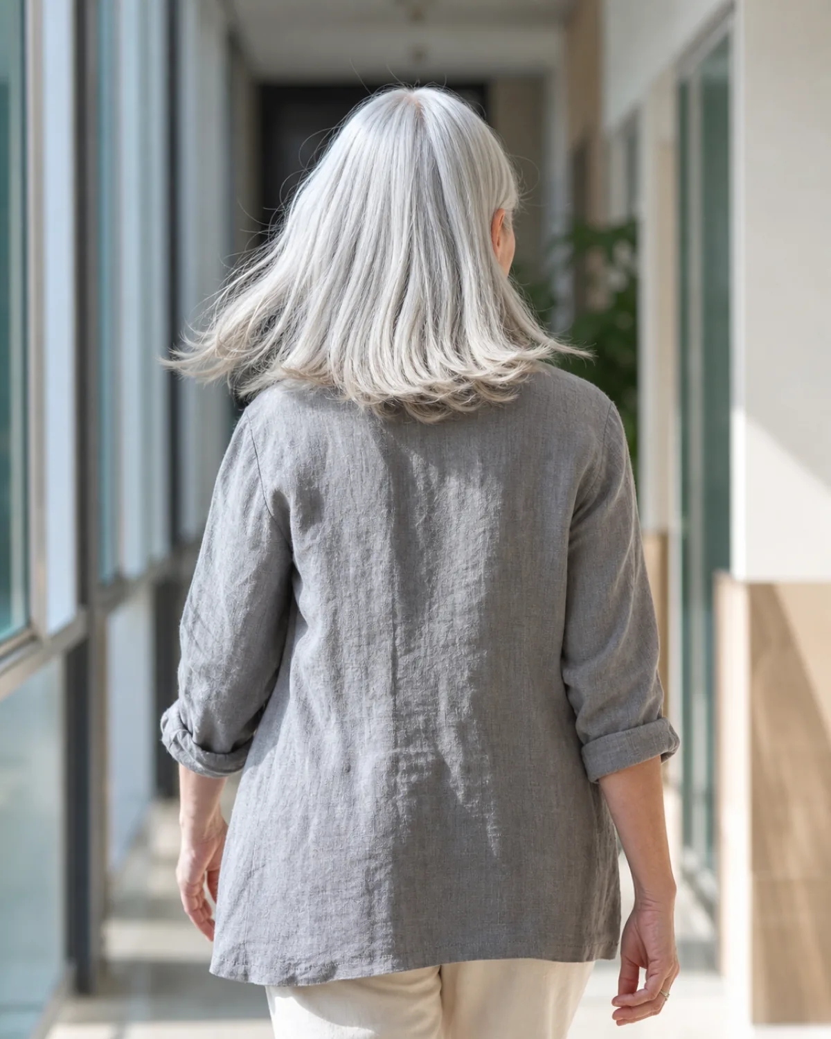 A mature woman with silver-white shoulder-length hair walking away through a bright hallway, shot from behind, showing a clean center part and no fringe pieces falling forward