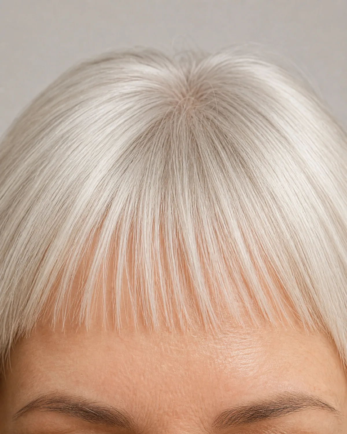 Extreme close-up of a mature woman with platinum silver hair showing a wispy micro-fringe cut short above the eyebrows, forehead skin visible through the thin strands