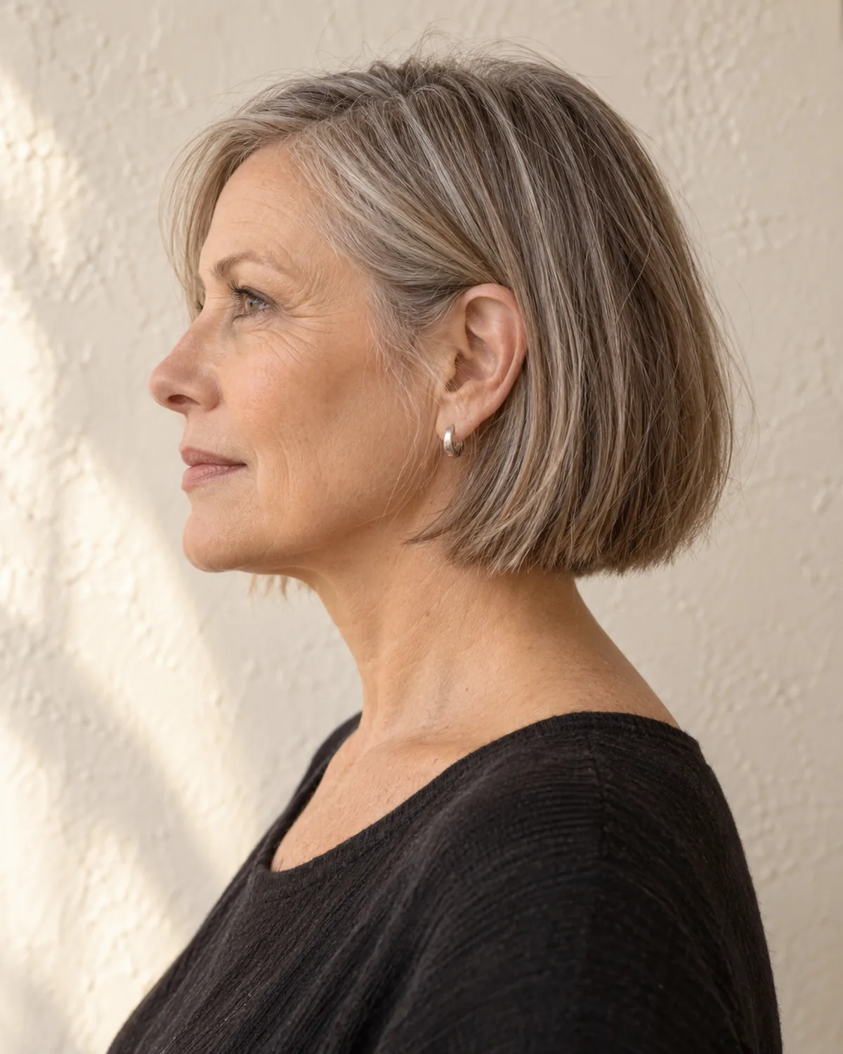 Mature woman with a chin-length bob in full side profile, showing the clean weight line ending precisely at the jawline against a warm textured wall