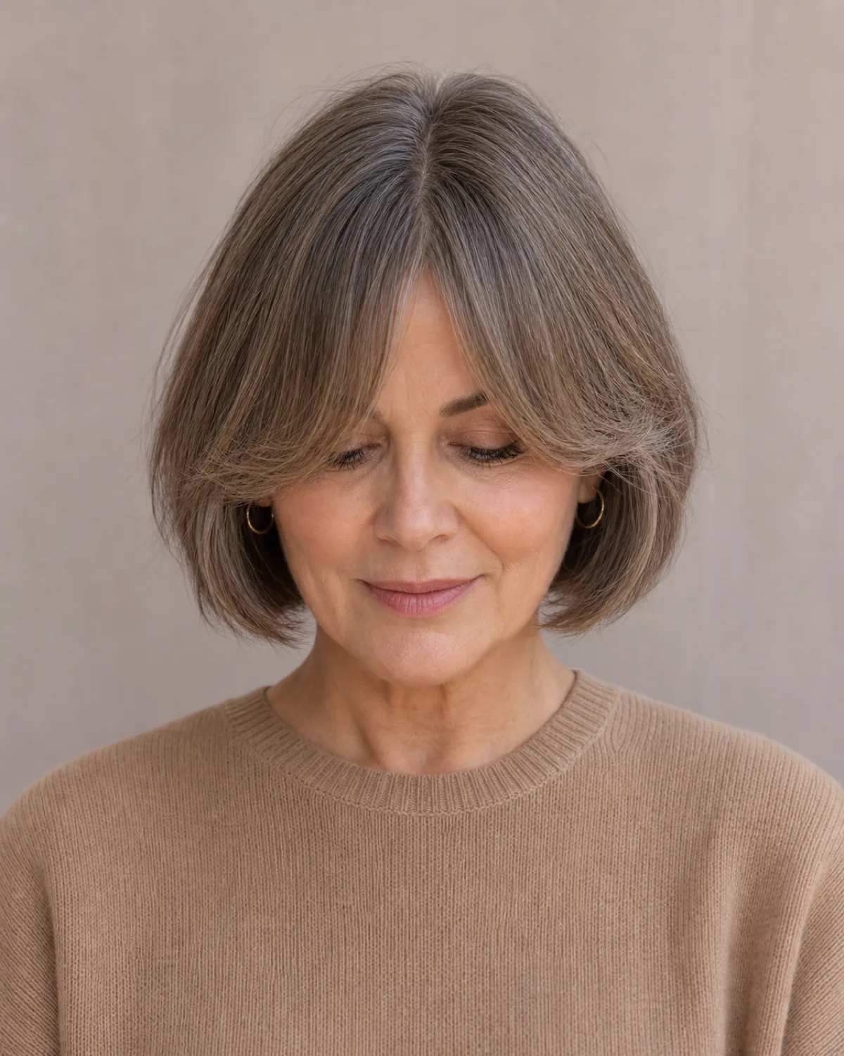 A front-facing view of a woman in a camel knit with a warm grey-brown bob and curtain bangs parting in the centre, the shortest point at the brow and the pieces sweeping toward the cheekbones