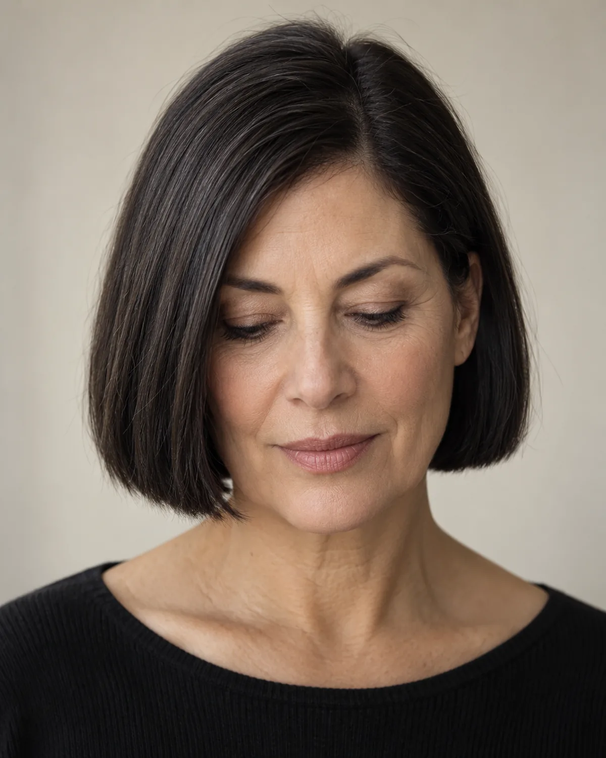 Front view of woman over 50 with dark blunt micro-bob ending exactly at the chin, eyes looking down, sharp hemline against neck
