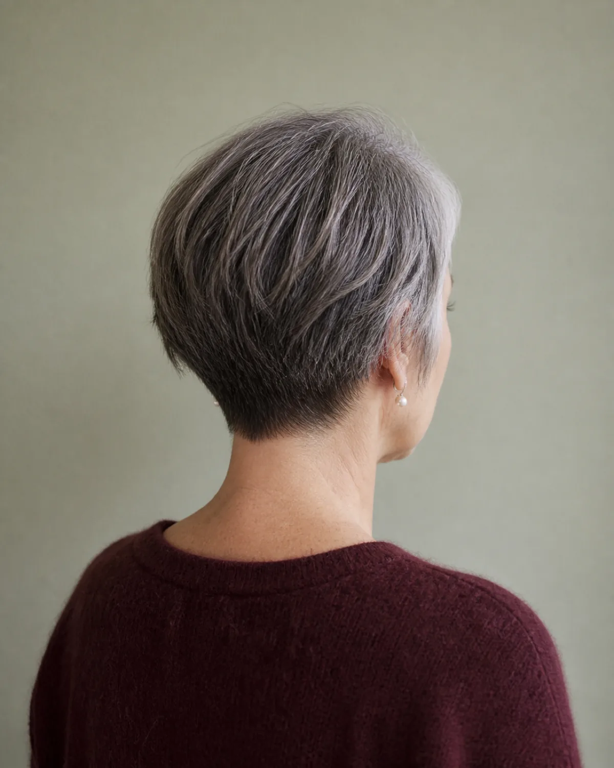 A back-three-quarter view of a woman with salt-and-pepper hair in a grown-out pixie, the nape showing a soft graduation that keeps the grow-out stage looking deliberate, against a pale sage wall