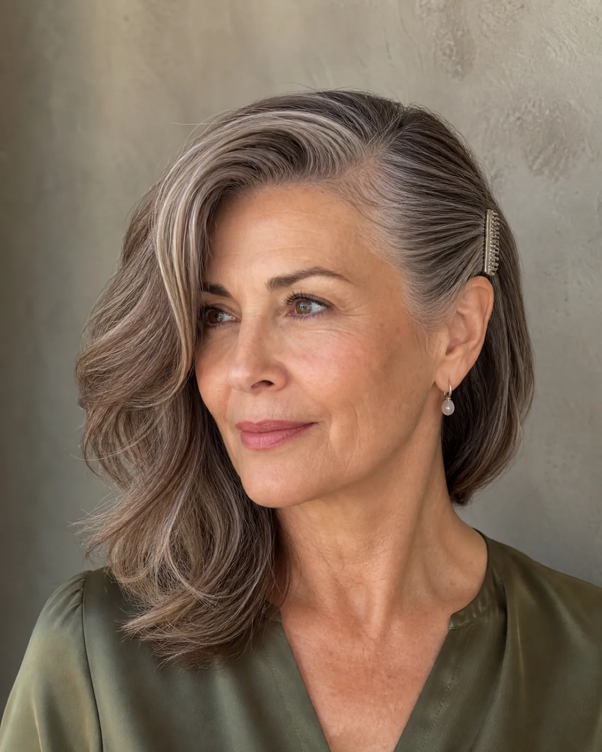 A woman in her early 60s turned three-quarters to camera left with a deep side part, wavy chestnut hair swept to one side, a decorative comb pinning the hair flat behind the opposite ear, warm light from above
