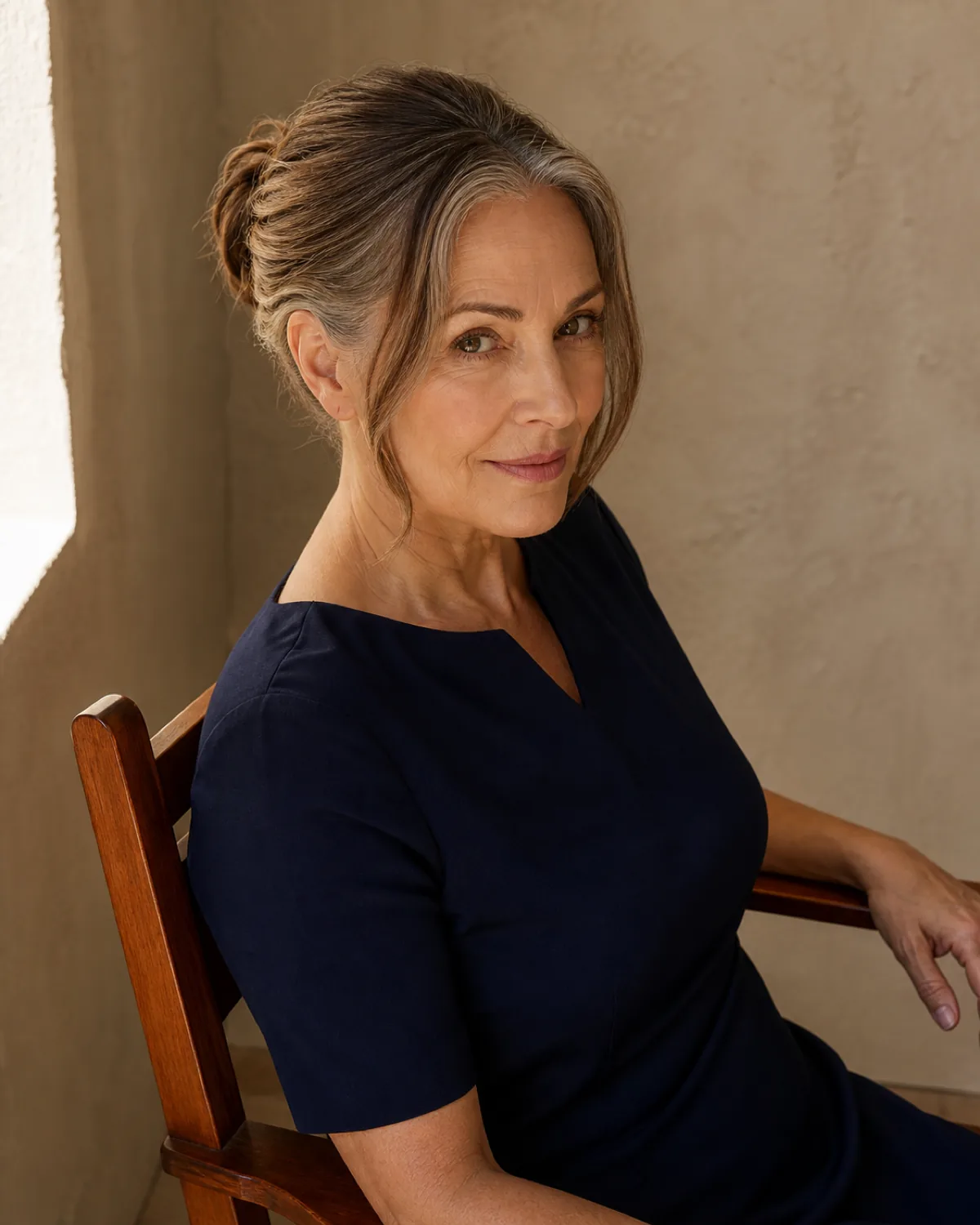A woman in her late 50s seen from slightly above and behind while seated, warm brown hair gathered in a loose bun at the mid-back with two soft face-framing pieces left out at the temples, one hand resting on her knee