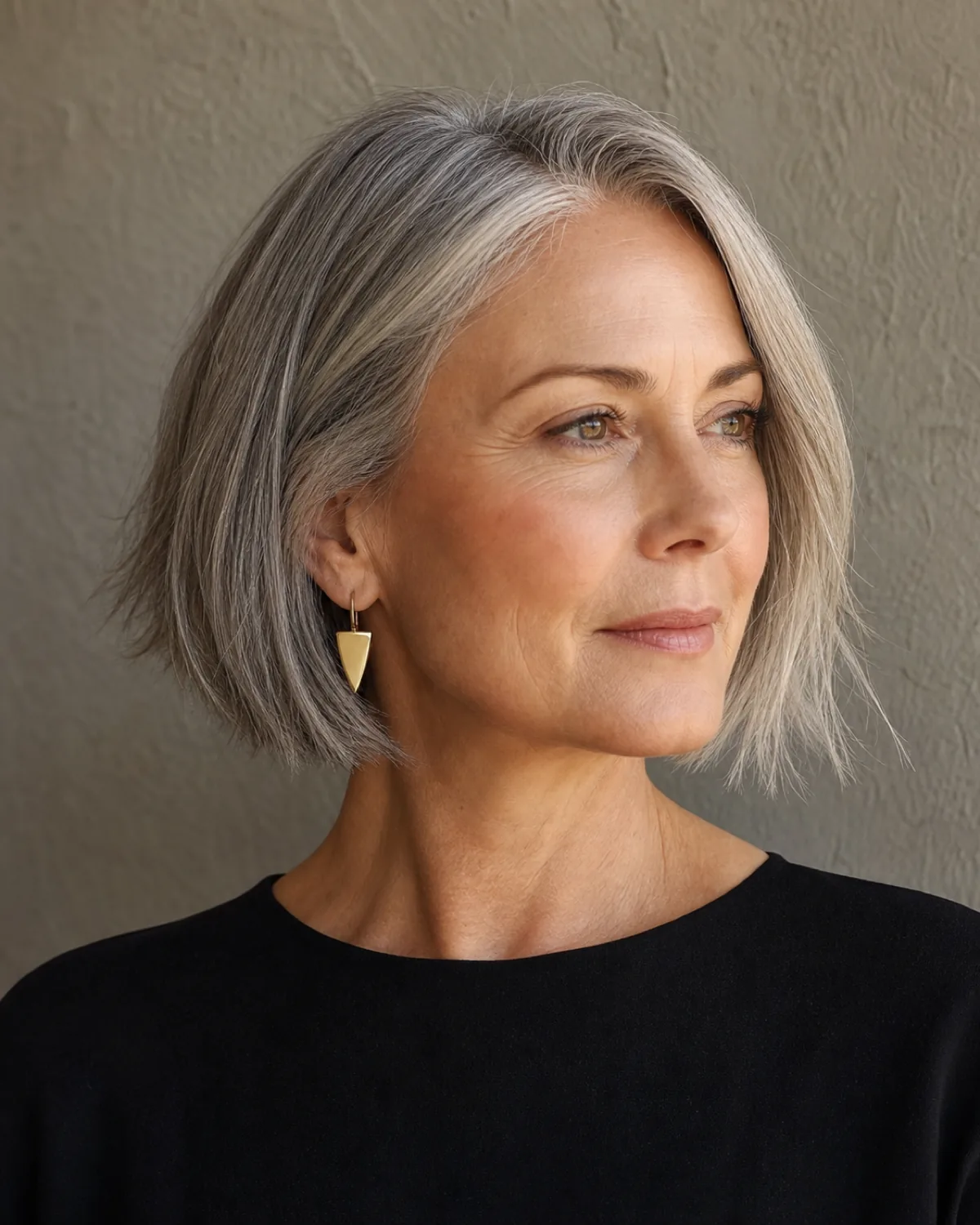 A woman in her early 60s turned three-quarters to camera left with a chin-length textured bob, point-cut ends separating into pieces, a gold drop earring visible on the near side, warm light from above