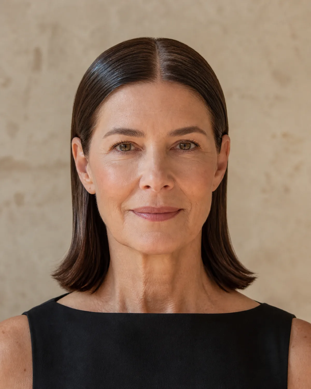 A woman in her late 50s facing the camera with dark brown hair in a centre-parted lob slicked flat with a glossy wet-look gel finish, the hair sitting close to the head, against a cool grey wall