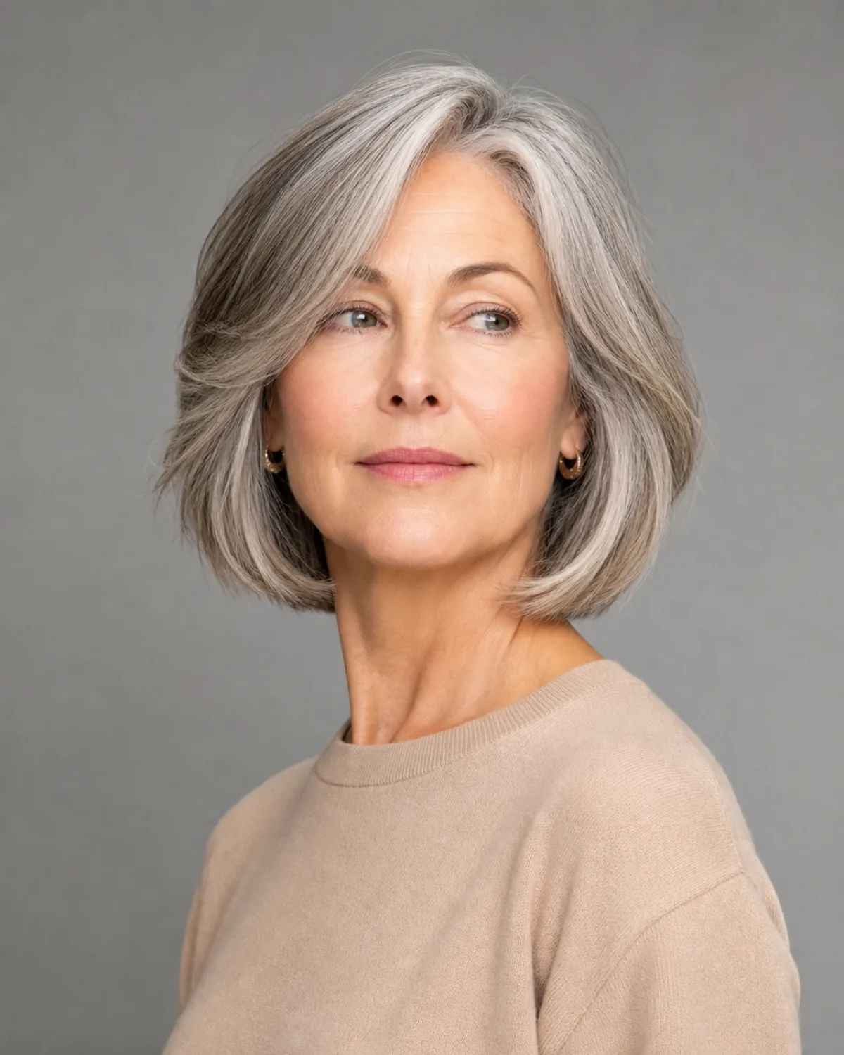 A woman in her early 60s with salt-and-pepper hair in a chin-length bob with soft face-framing layers, turned three-quarters to camera left with chin slightly lifted, the tapered ends breaking the horizontal line
