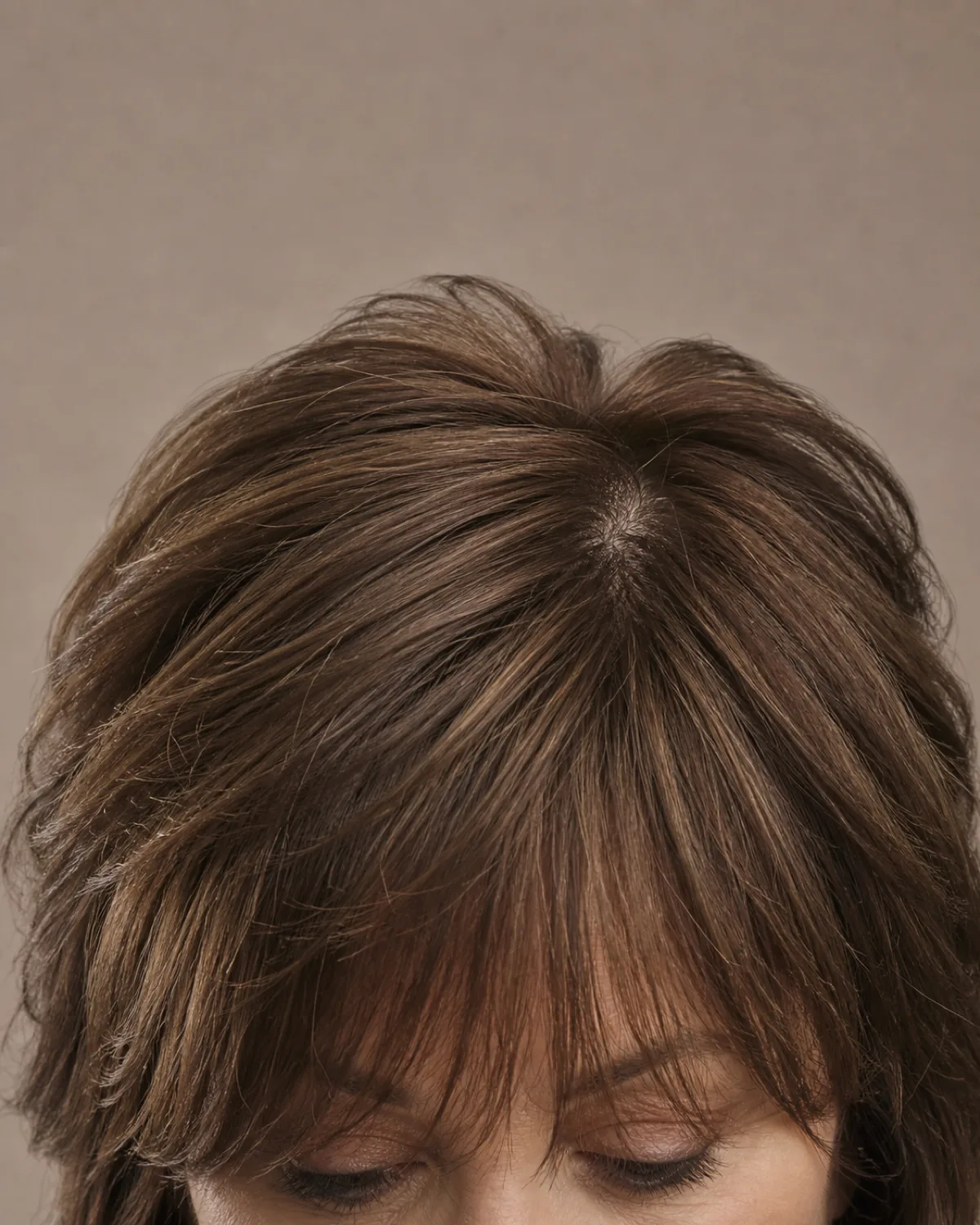 Close crop on the fringe and crown of a woman with warm brown hair in a medium-length shag, layered movement radiating from the centre part, eyes below the frame edge