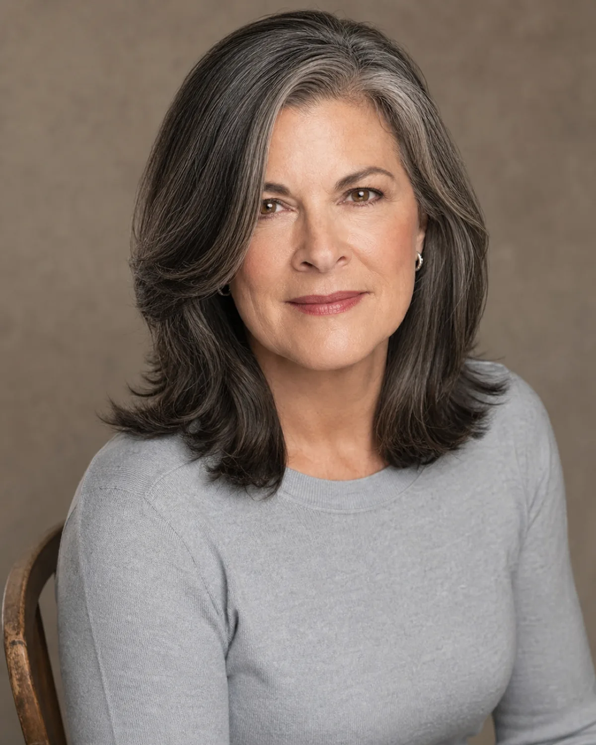 A woman in her late 50s with dark hair with silver threads at shoulder length, seated with layers falling below the cheekbone, softening the jawline