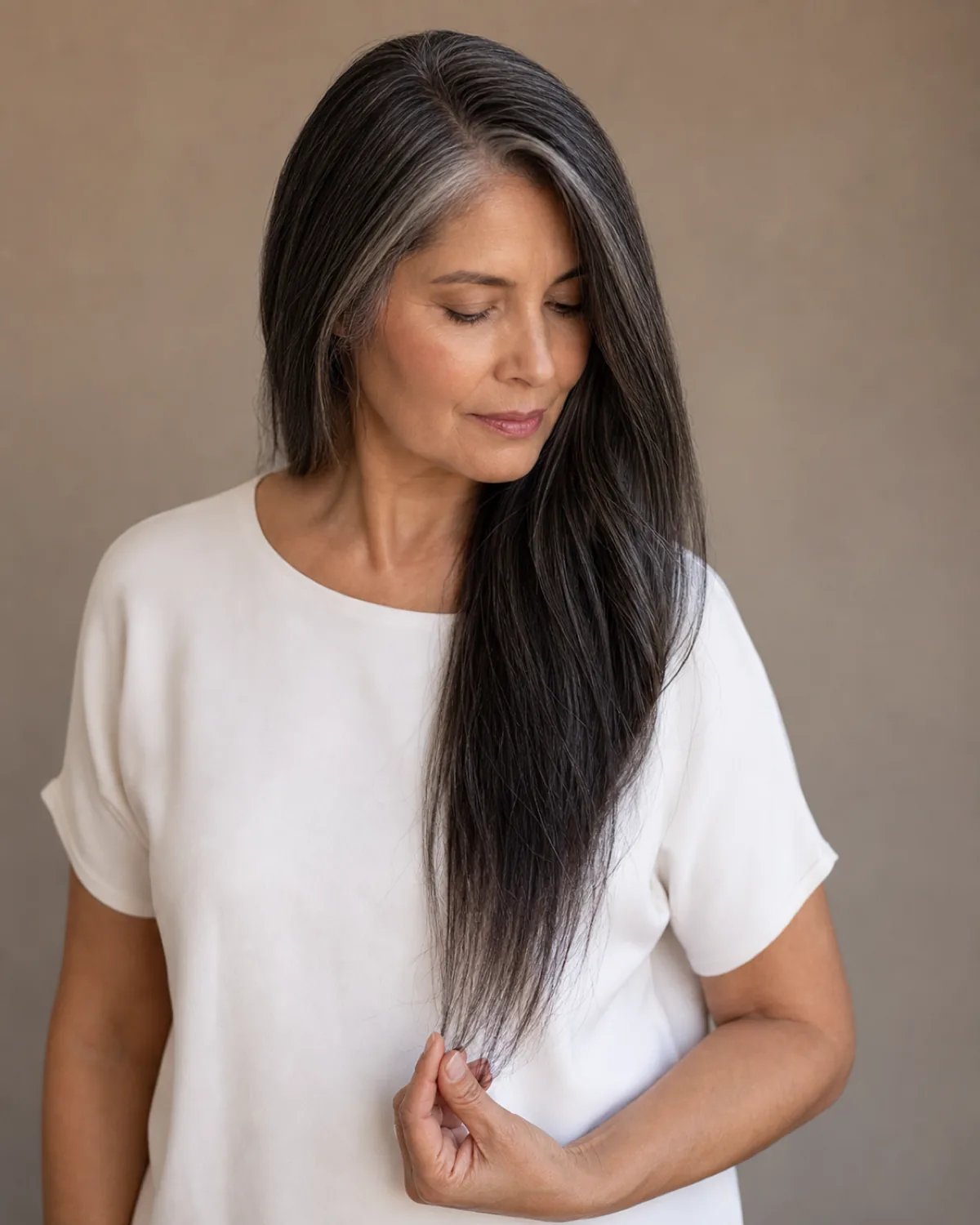 A woman in her late 50s with long dark hair with grey strands, looking down at the ends she holds between her fingers, the density visibly thinner in the lower portion