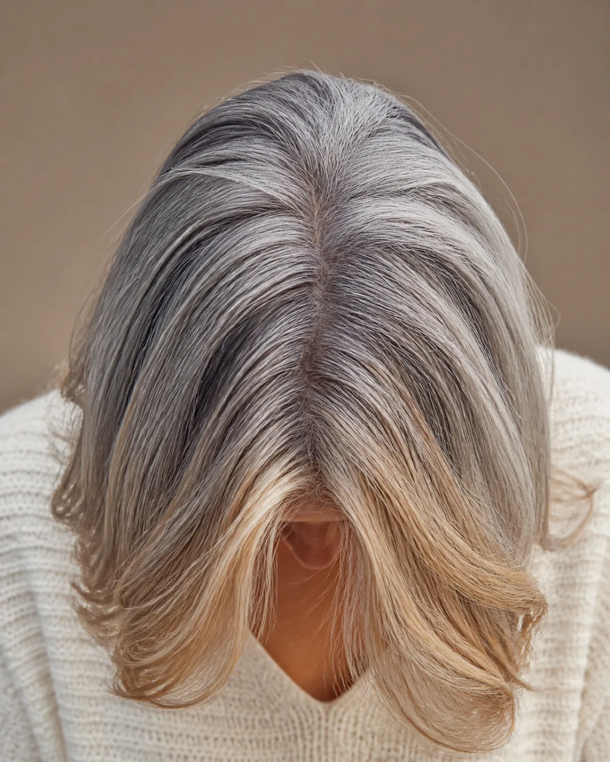Crown-forward view of a woman with mostly silver hair and warm honey-blonde face-framing highlights at the temples, cream sweater, face not visible