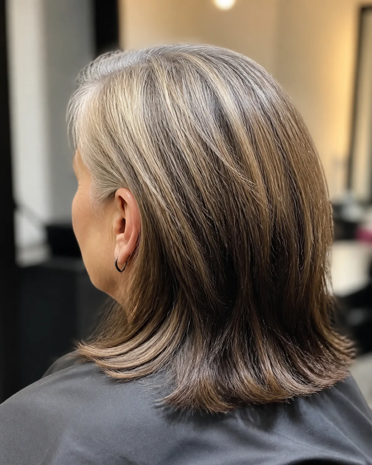 Back three-quarter view of a woman with highlights placed near the gray root band to soften the demarcation, salon cape, shoulder-length hair