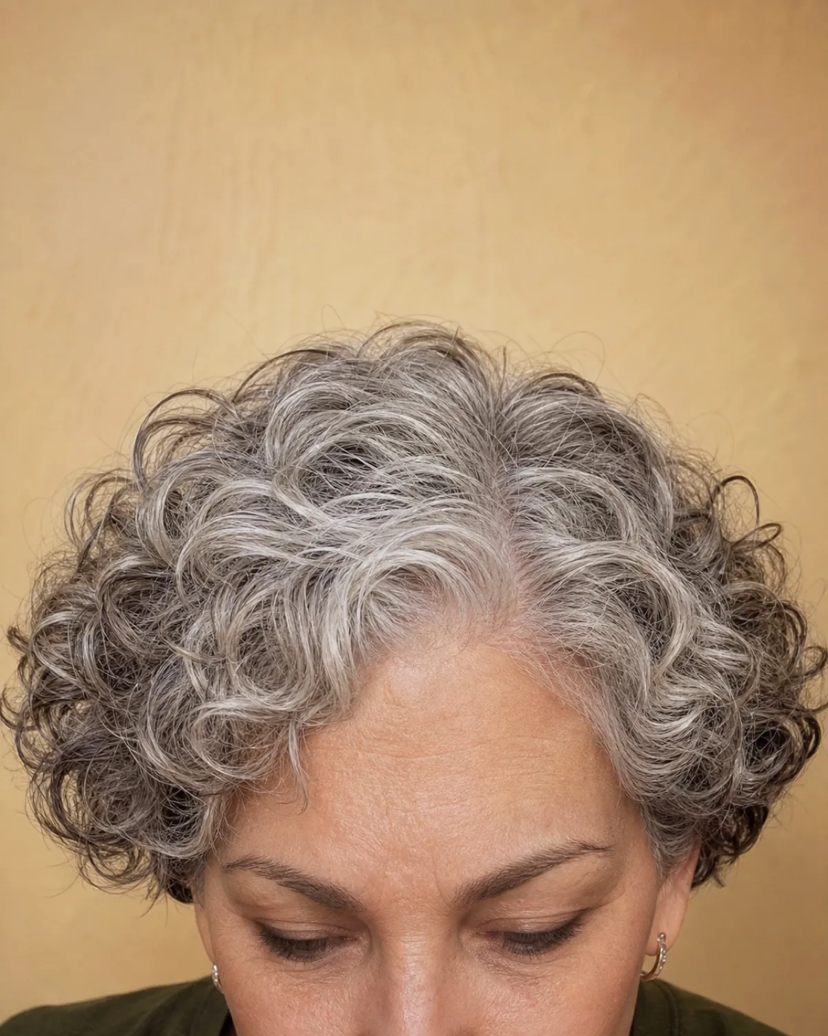 Top-down close view of an ear-length silver-grey curly bob showing dense springy curls hugging the head from fringe to crown, brows and downcast eyes visible at the frame edge
