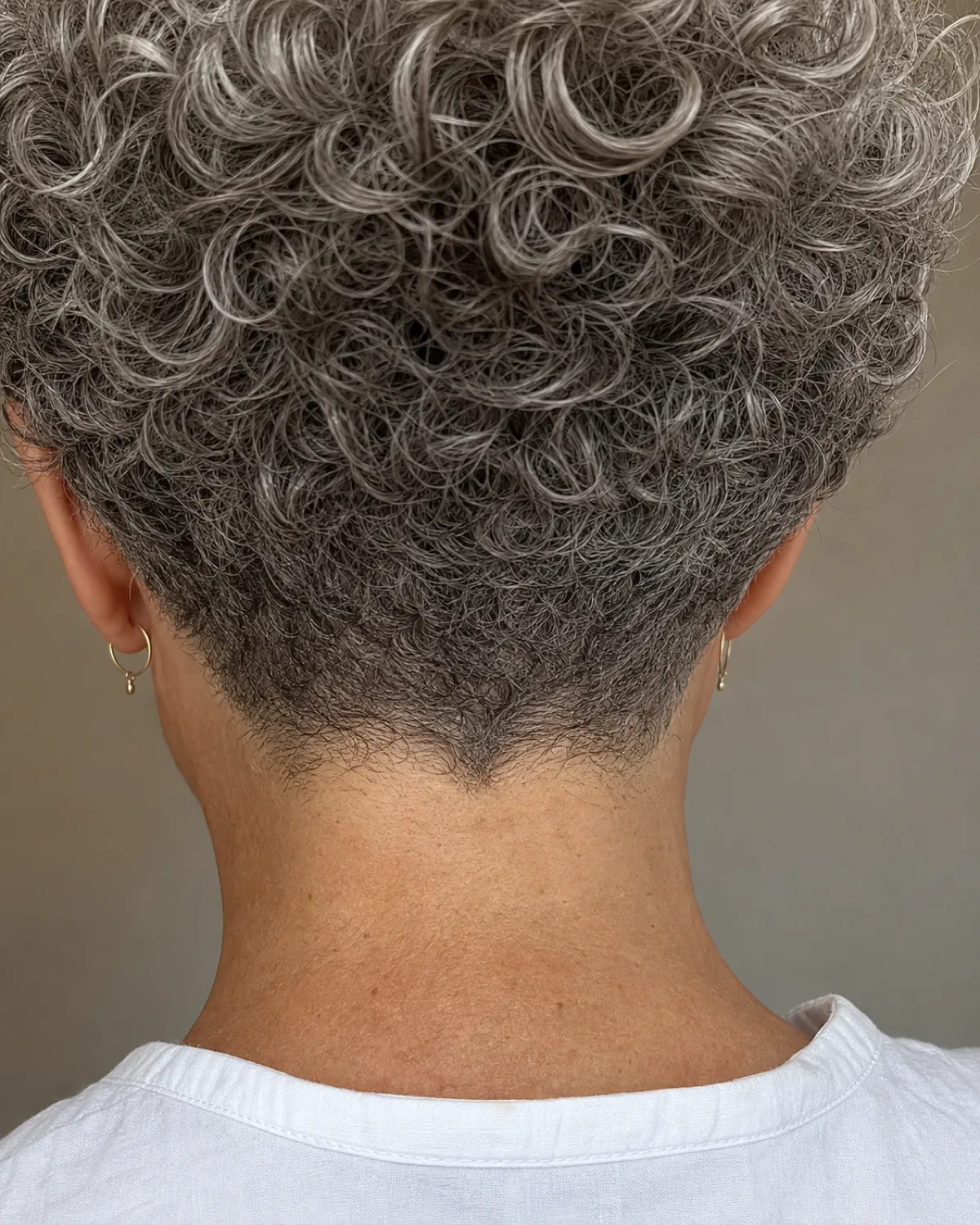 Close-up of the back of a silver-grey curly bob tapered to a tight point at the nape, curls graduating from full at the crown to short and close at the hairline, small gold hoop earrings