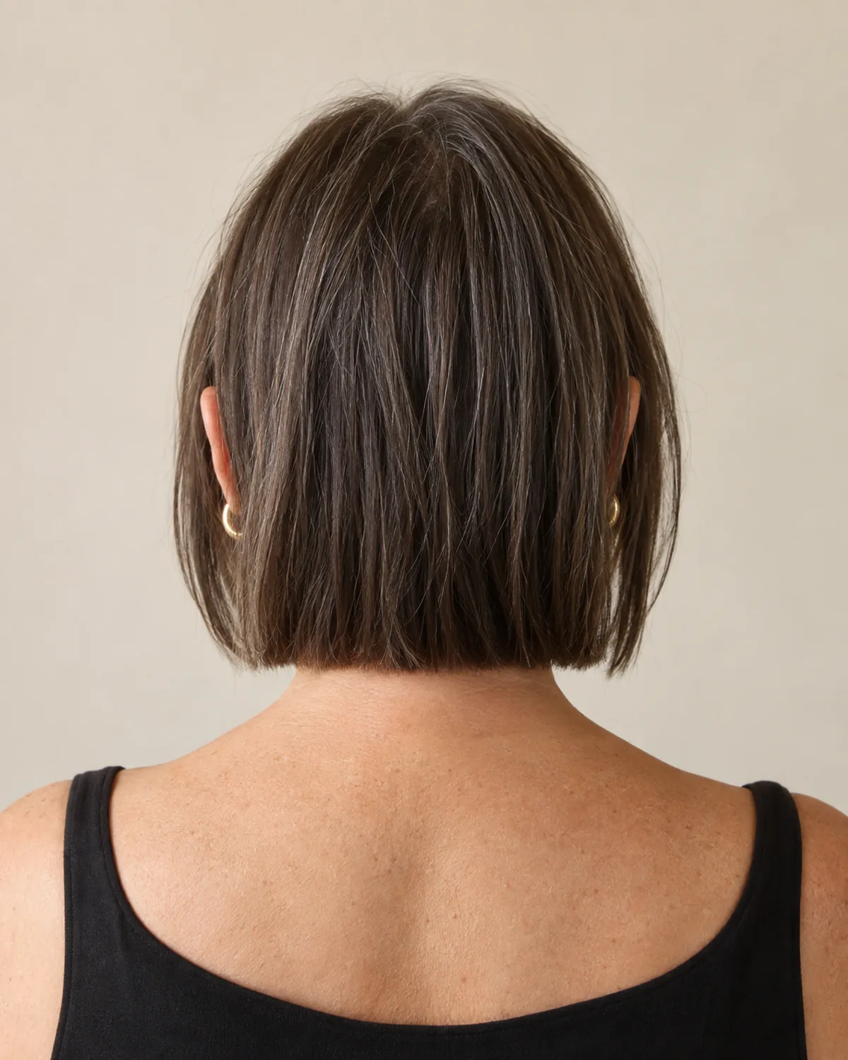 Back view of a mature woman with a blunt horizontal perimeter at the nape, dark brown hair with scattered silver, the clean back line visible
