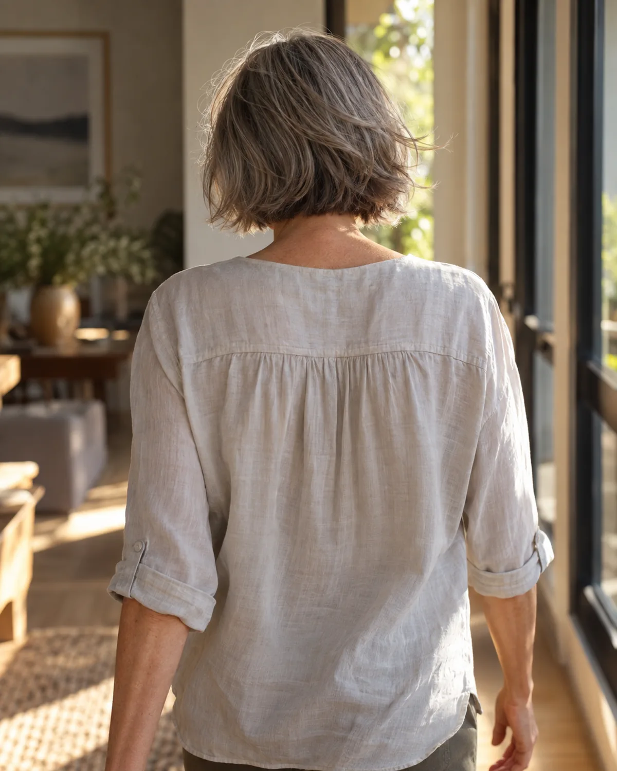 Mature woman walking away from camera, jaw-length wispy layers in gentle motion, shot from behind at eye level in warm afternoon light