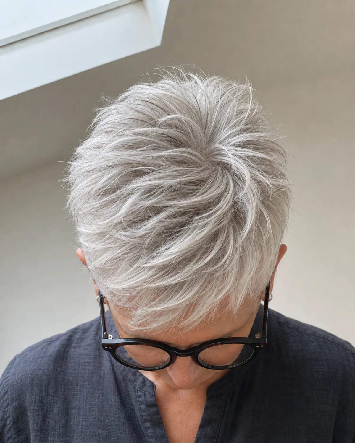 A woman in her early 60s with a bright silver-white textured pixie cut viewed from above, head tilted forward to show piecey separated crown layers with volume and lift, bold round dark-framed glasses visible below