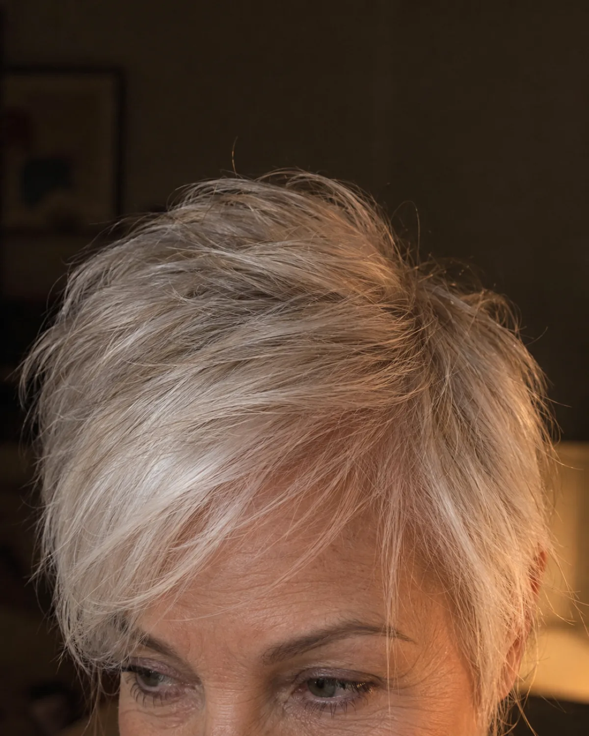 Close crop of a mature woman with fine platinum-white hair in a wispy pixie, longer textured pieces at the top and fine individual wisps at the temples, eyes at the bottom edge of the frame