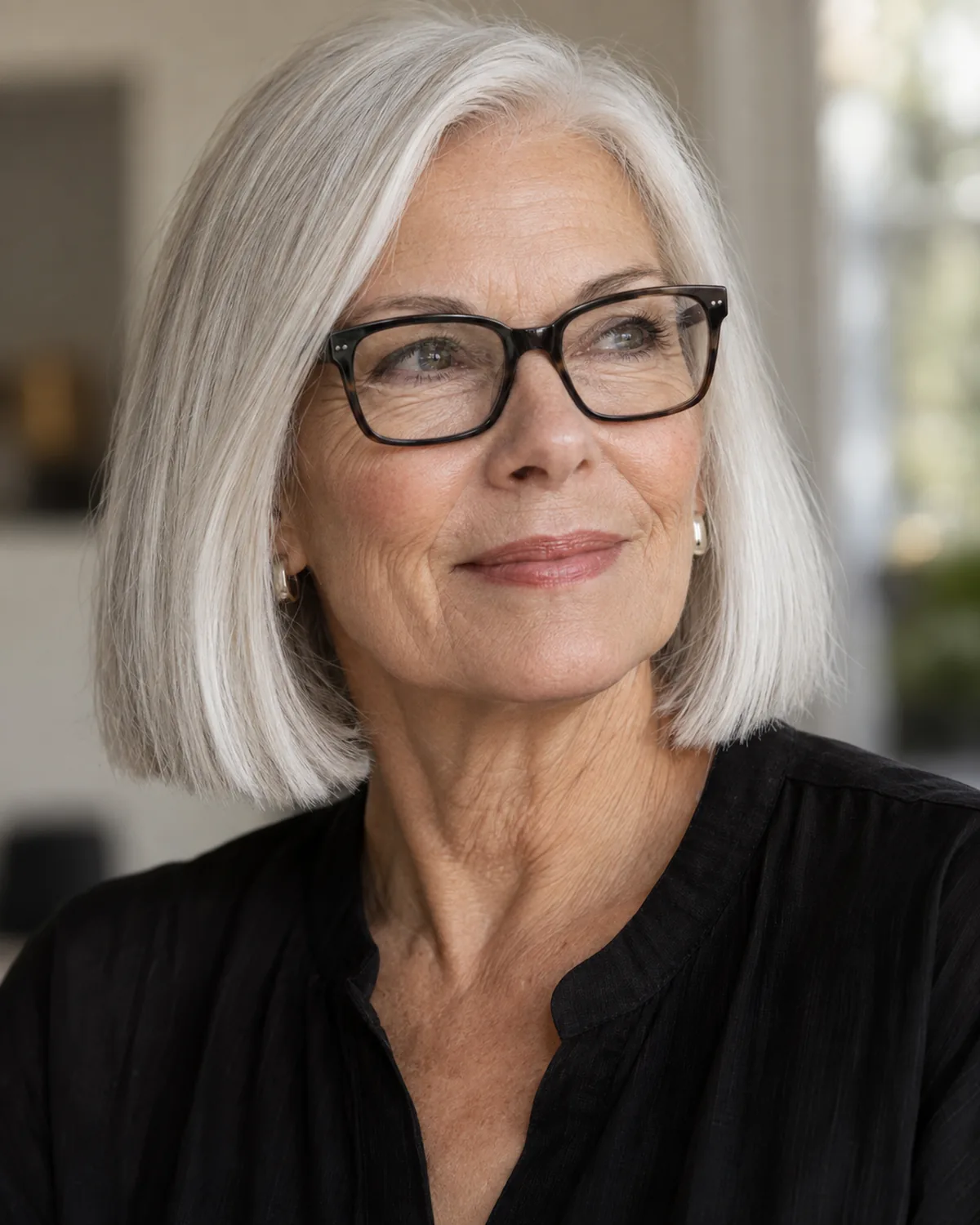 A woman over 60 with fully white-silver hair in a one-length bob at chin level, close crop showing blunt ends, dark rectangular glasses creating strong contrast with the white hair, soft neutral background