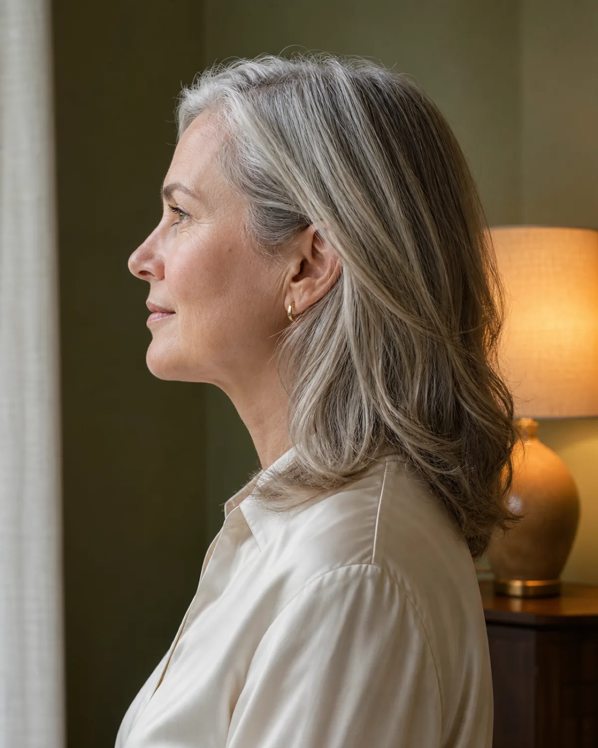 A woman in full profile from the side, warm silver shoulder-length hair with soft layers, in a cream silk blouse against a sage-green wall with a warm table lamp behind her