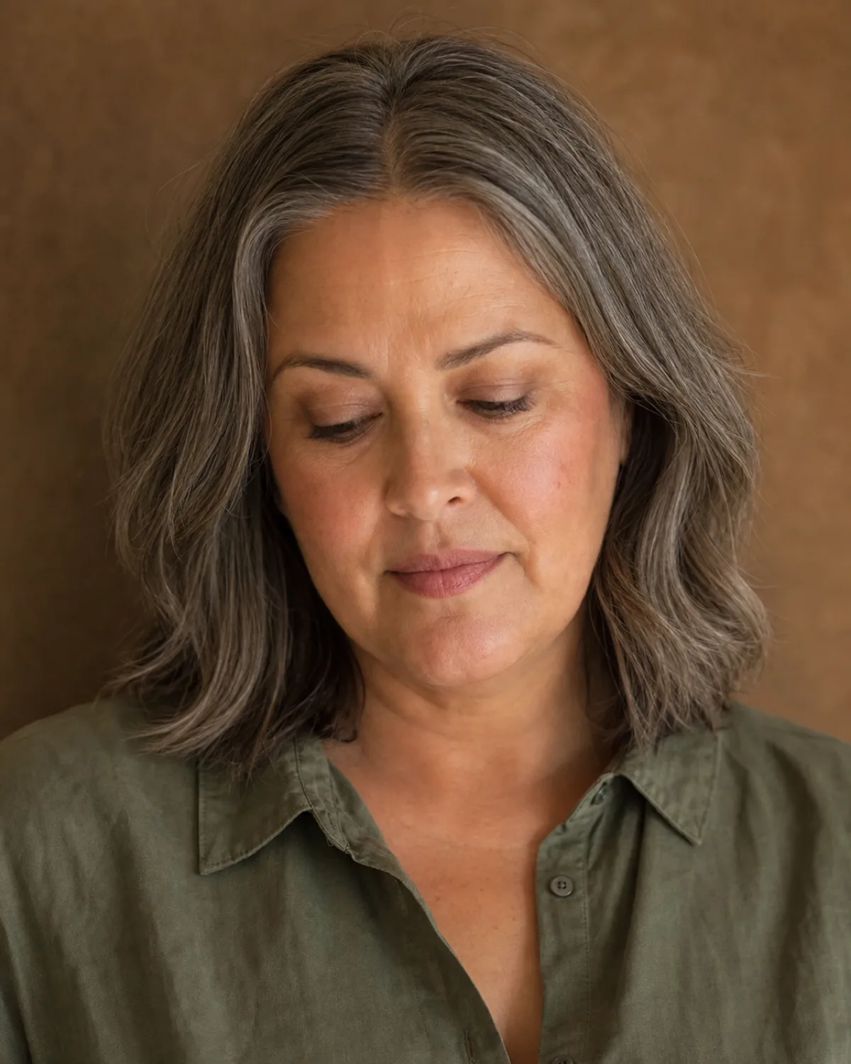 A woman with salt-and-pepper collarbone-length hair facing the camera with eyes downcast, her face shape ambiguous between round and heart, wearing an olive linen shirt against a clay wall