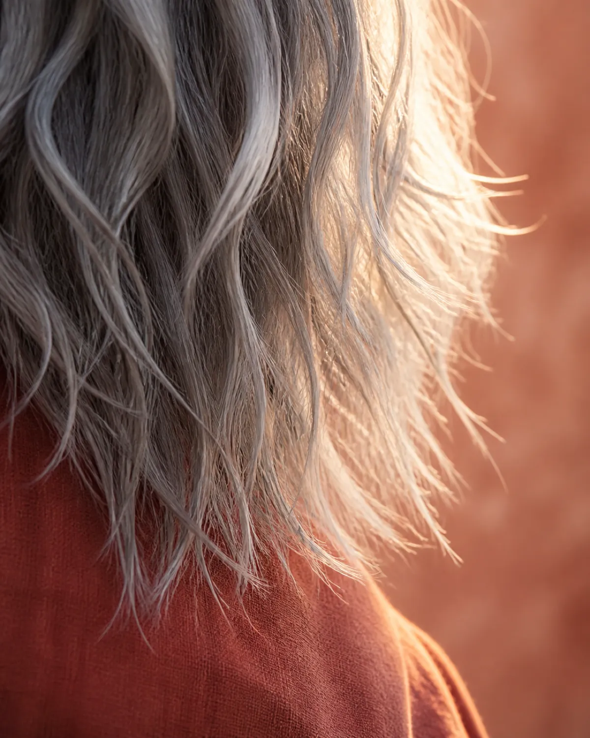Extreme close-up of wavy silver-grey razor-cut hair ends backlit by warm daylight, individual strands ending at different lengths in a choppy textured finish