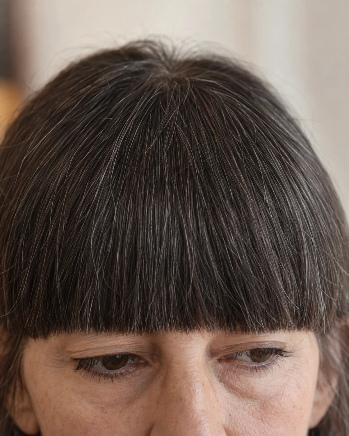 Close crop on the fringe and brow line of a mature woman showing thick blunt bangs cut straight across at the eyebrow