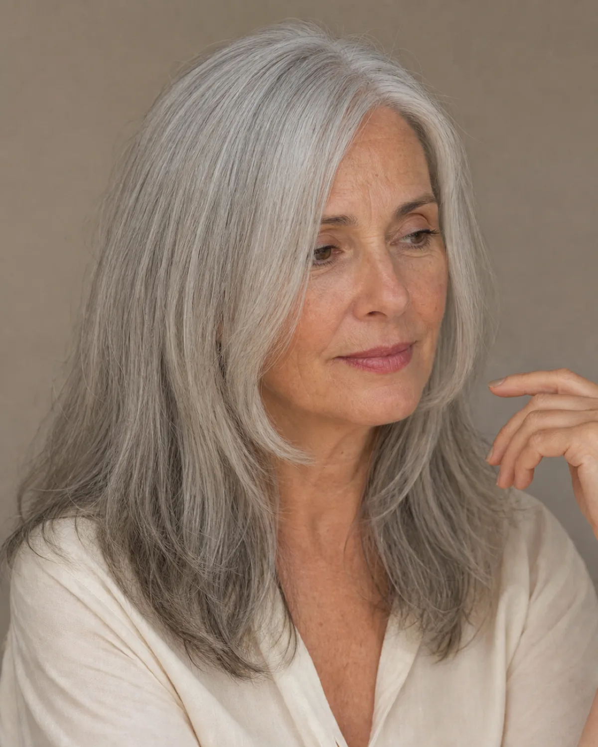 Three-quarter view of a woman in her early 60s with long silver-grey hair and two shorter face-framing pieces falling along each side of the jaw, wearing a cream blouse against a taupe background