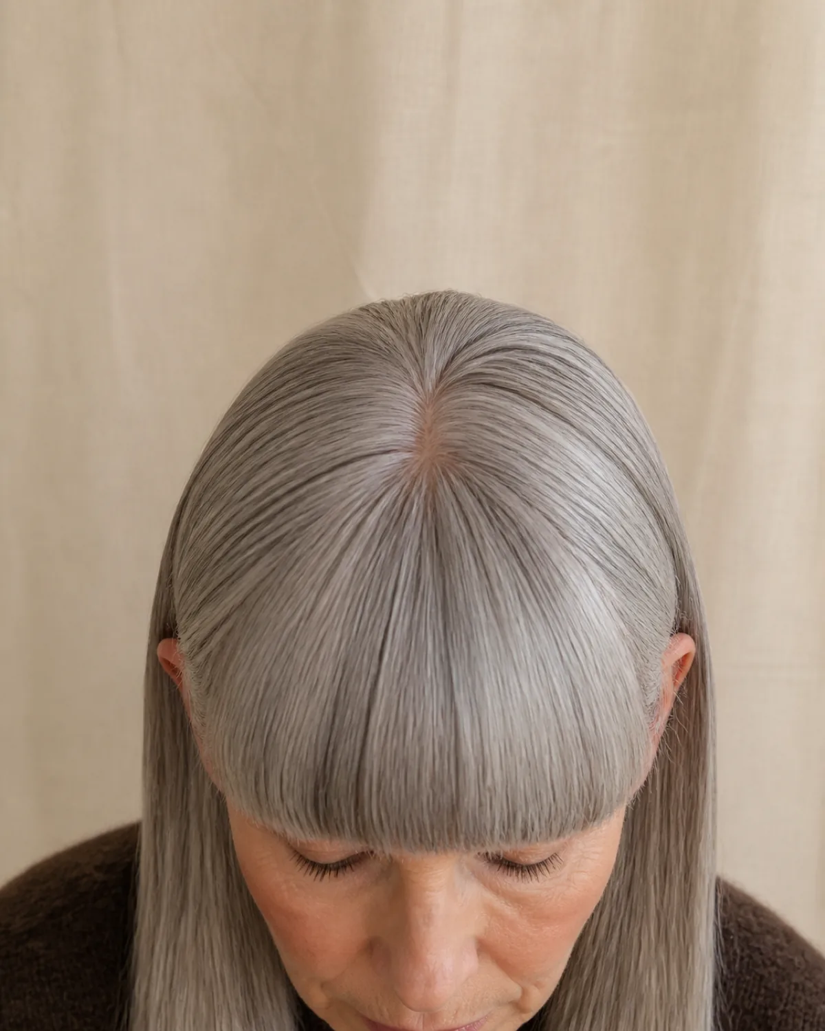 Close crop showing the fringe and brow line of a woman with thick straight silver-grey hair and a dense blunt fringe cut in a single straight line just above the eyebrows, wearing a dark chocolate brown knit against a beige linen background