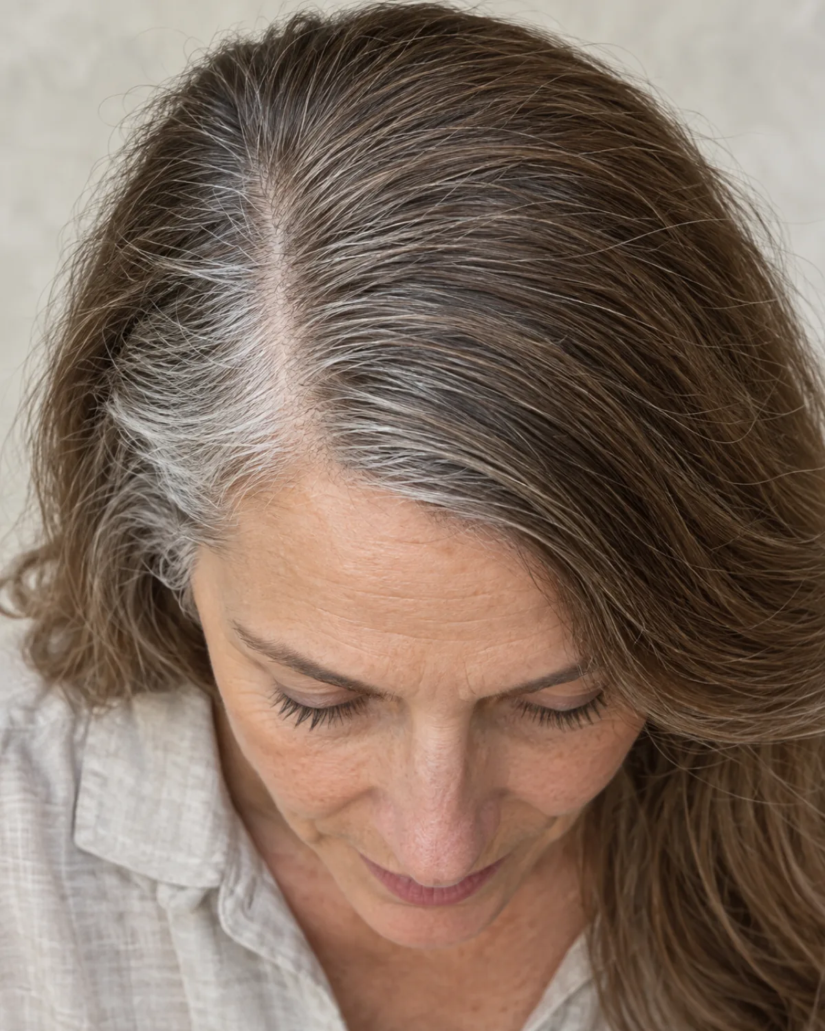 Top-down close crop showing deep side part on fine hair, part line visible with grey at temples, asymmetric volume stacking more hair to one side, oatmeal linen shirt