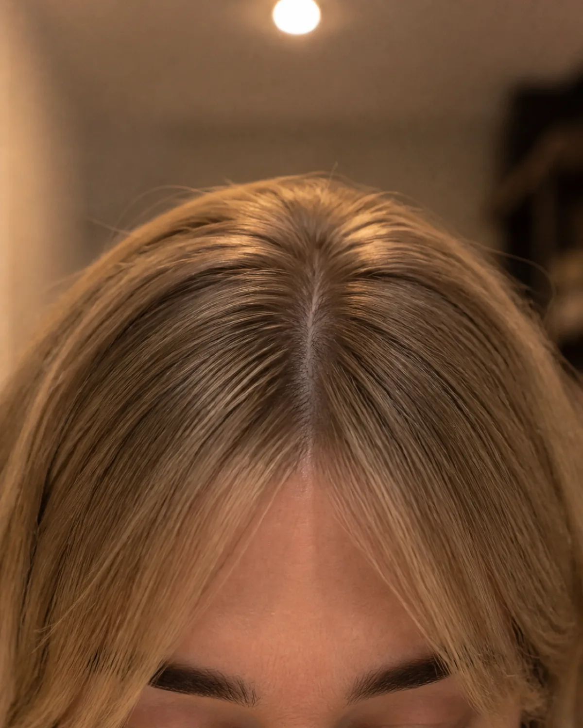 Close crop on the fringe and brow line of a mature woman with fine dark-blonde hair, showing a narrow curtain fringe parted cleanly in the centre with warm overhead lamplight, eyes just below the frame edge