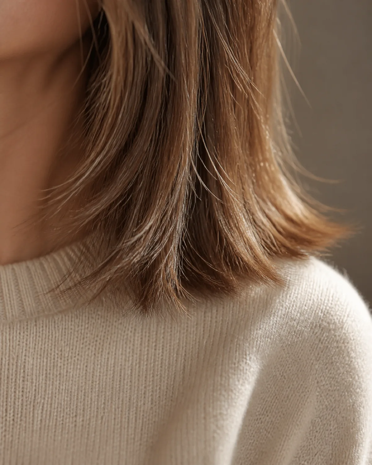 Close crop on the ends of a mature woman with fine light-brown hair in a razored collarbone cut, showing feathered and separated tips where individual strands splay slightly at the cutting edge against a cream knit sweater