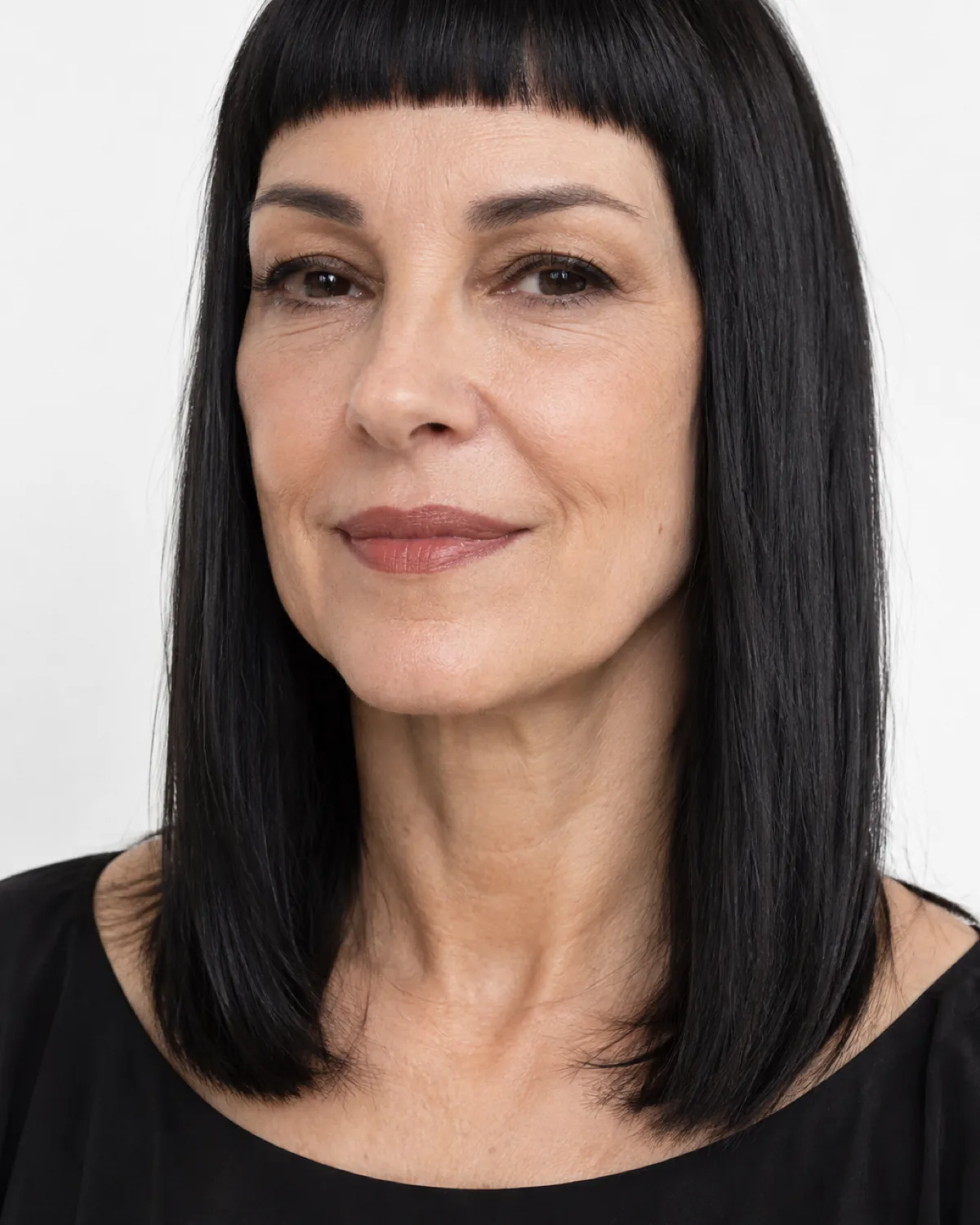 Woman over 50 with jet black sleek midi hair and baby bangs cut in a short horizontal line above mid-forehead