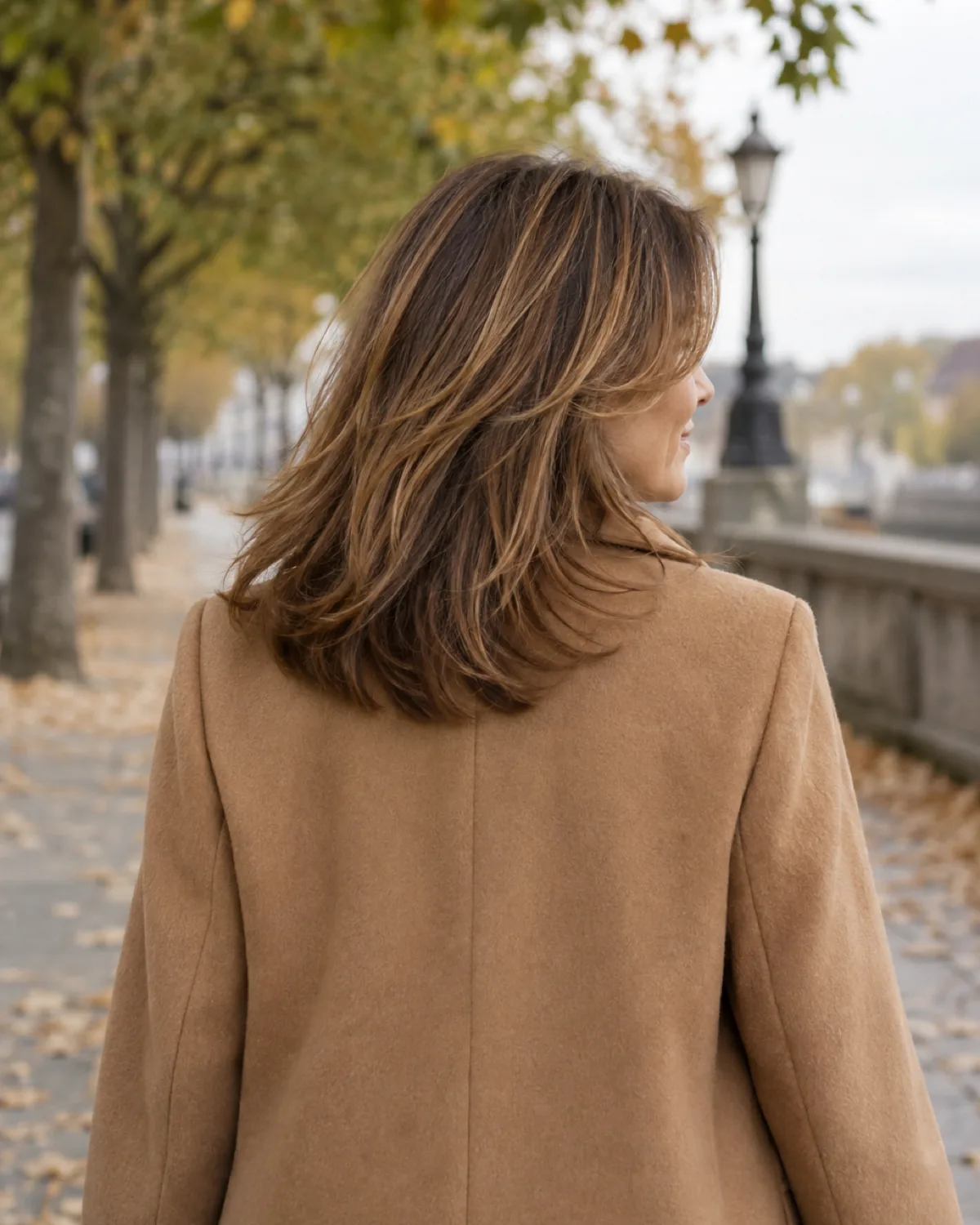 Woman over 50 walking away with medium brown hair in motion, long bangs tapering into face-framing layers, wearing a camel coat