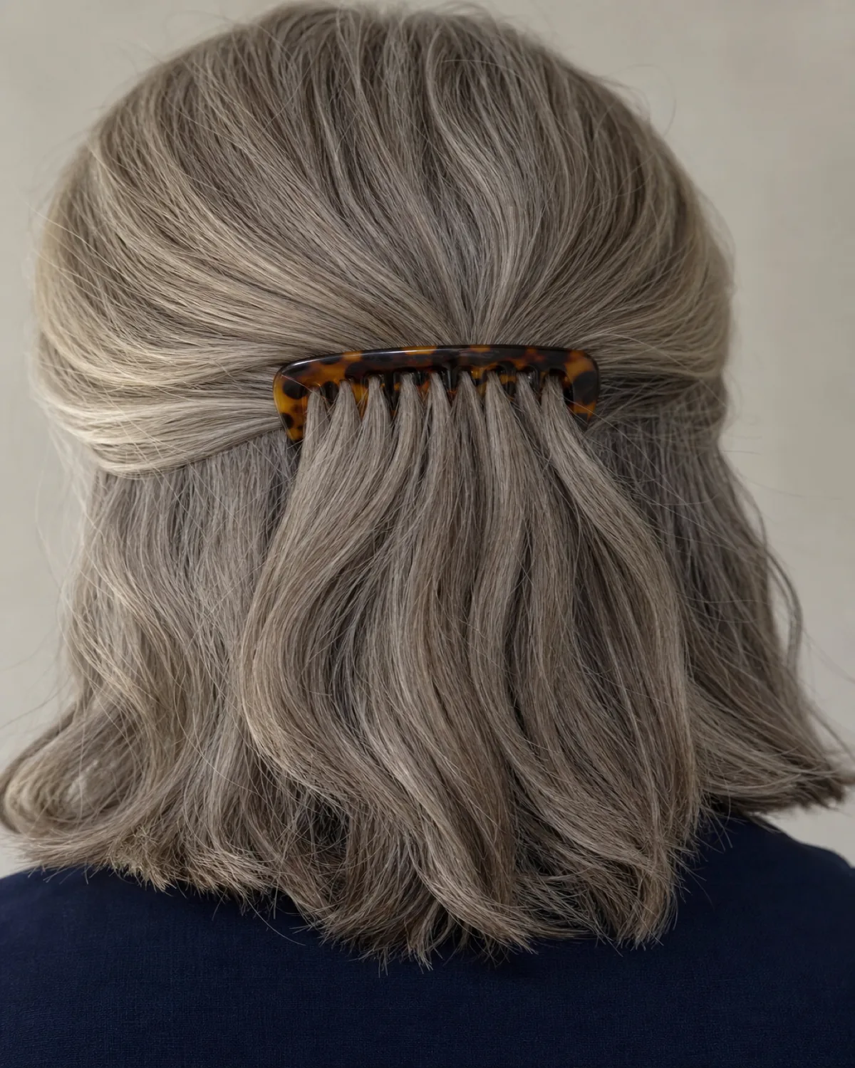 Close crop of the back of a woman's head with warm grey hair, a tortoiseshell decorative comb inserted horizontally just below the crown, holding the upper section gathered while the lower section hangs down past the nape, wearing a dark navy collar