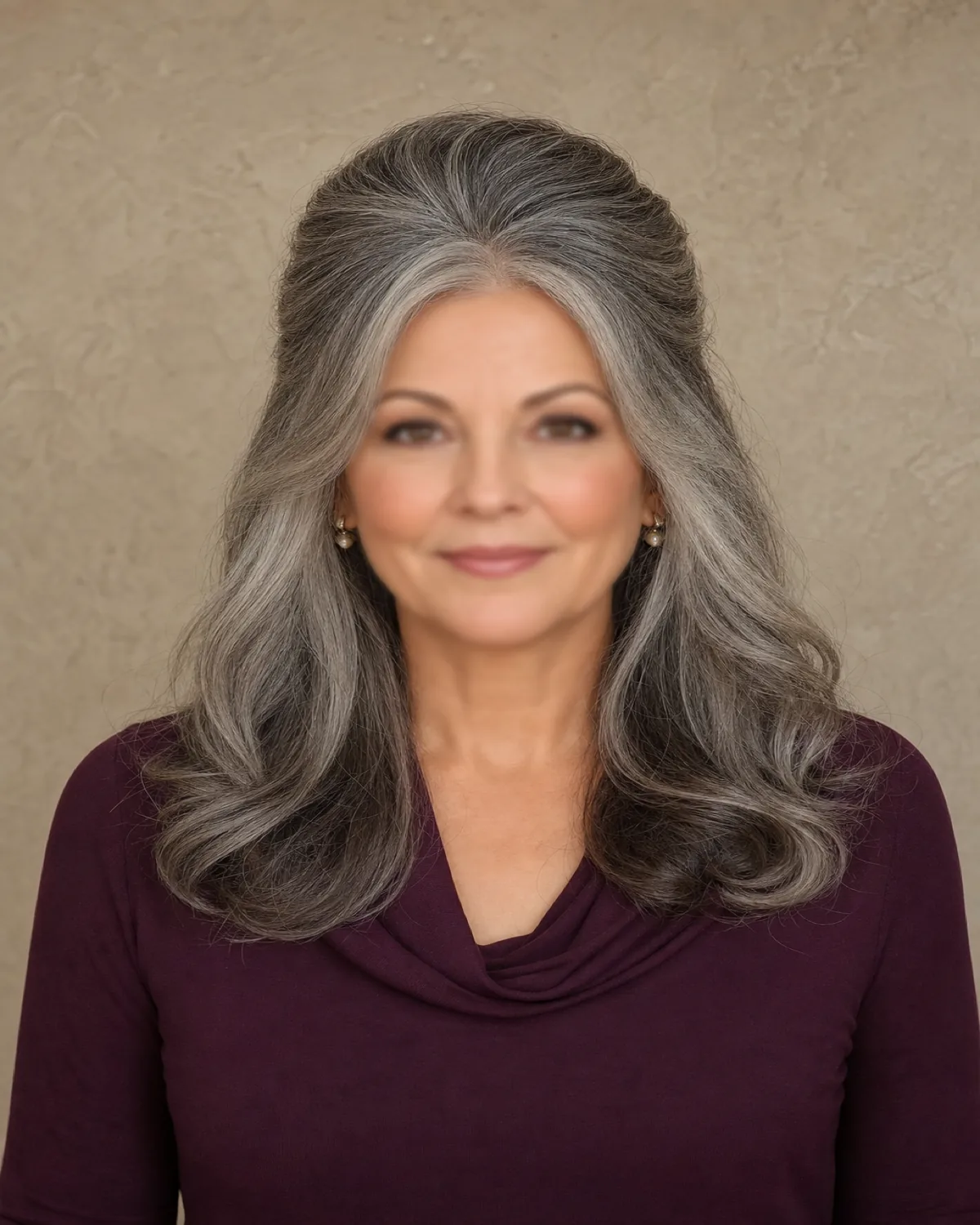 A front-facing view of a woman with salt-and-pepper hair swept back from the forehead with visible root lift at the crown, the lower half falling in soft curled ends past her shoulders, wearing a deep plum cowl-neck top against a warm taupe plaster wall