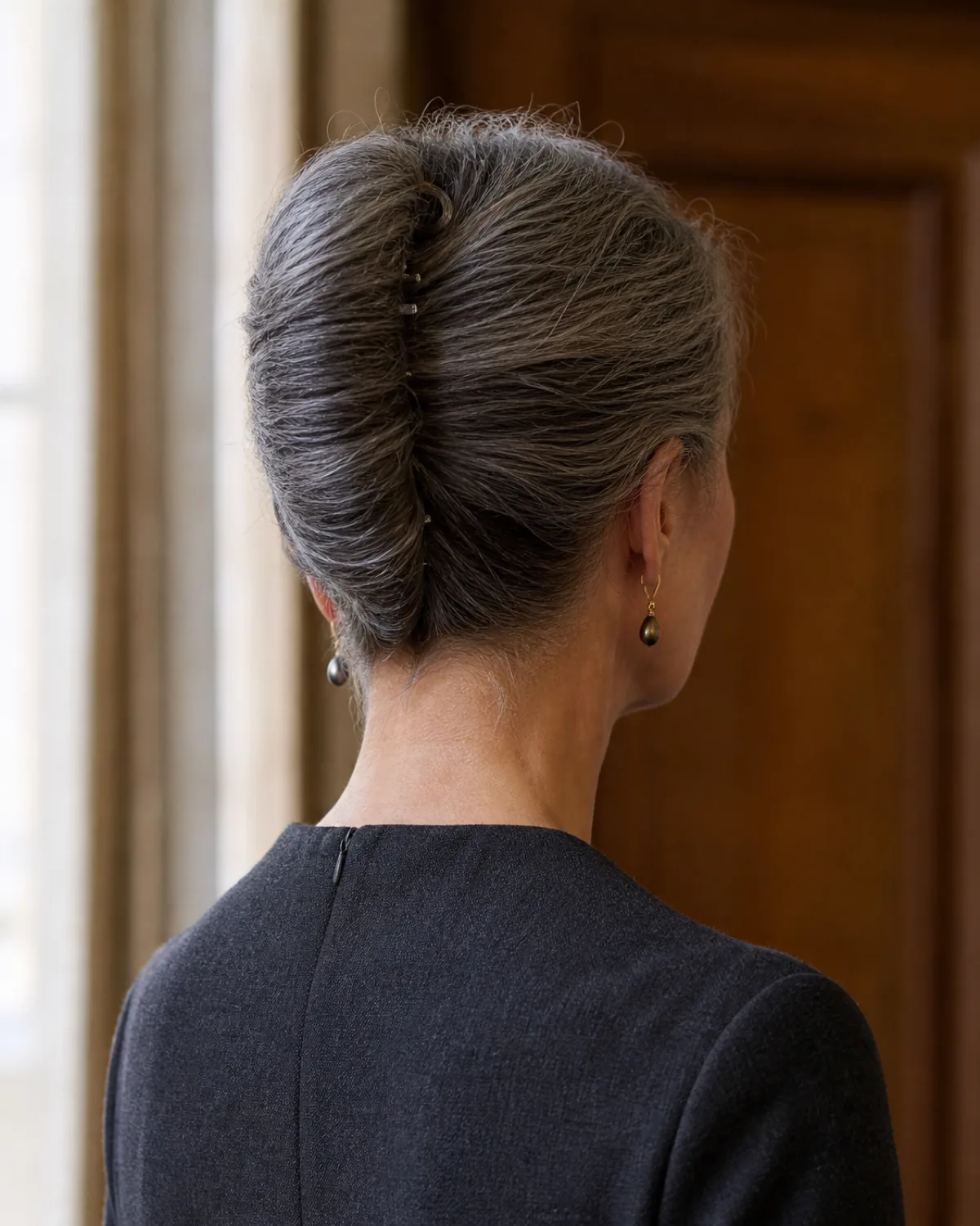 Back view of a woman with salt-and-pepper hair in a classic French twist, the vertical roll running from nape to crown with visible pin placement along the seam, wearing a charcoal wool dress