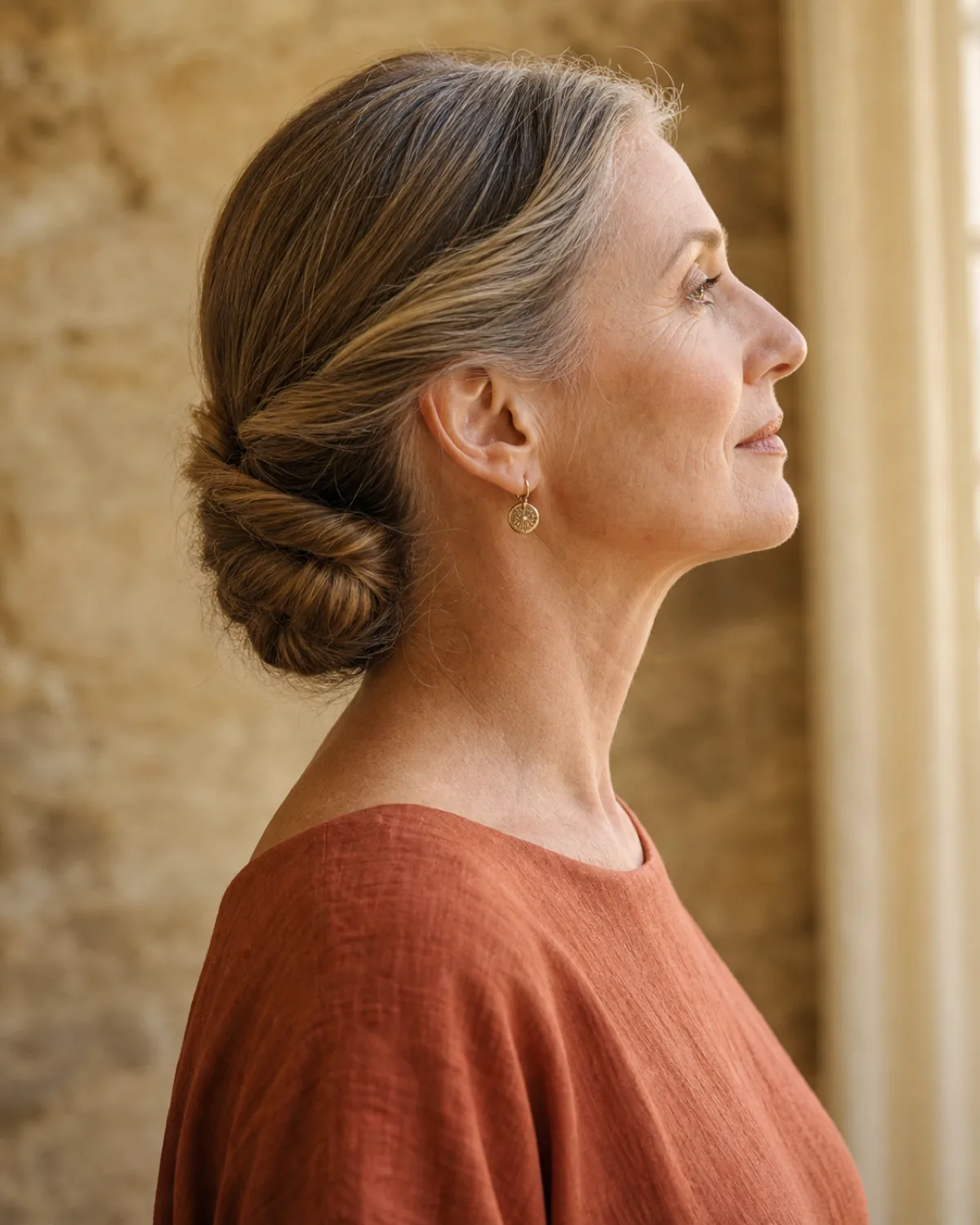 Side profile of a woman with dark-blonde silver-threaded hair in a double-twisted low roll at the nape, two twists crossed and interlocked forming a flat roll, wearing a terracotta linen dress