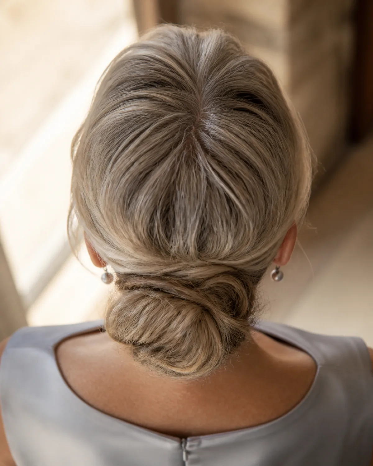 Top-down view of a woman's head showing gentle crown lift above a loose low bun at the nape, warm grey hair with a single central part, pearl-grey silk dress visible
