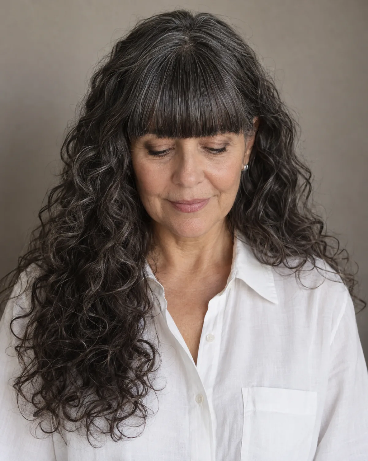 Front-facing mature woman with eyes downcast, long curly hair with straight-across blunt bangs at brow level