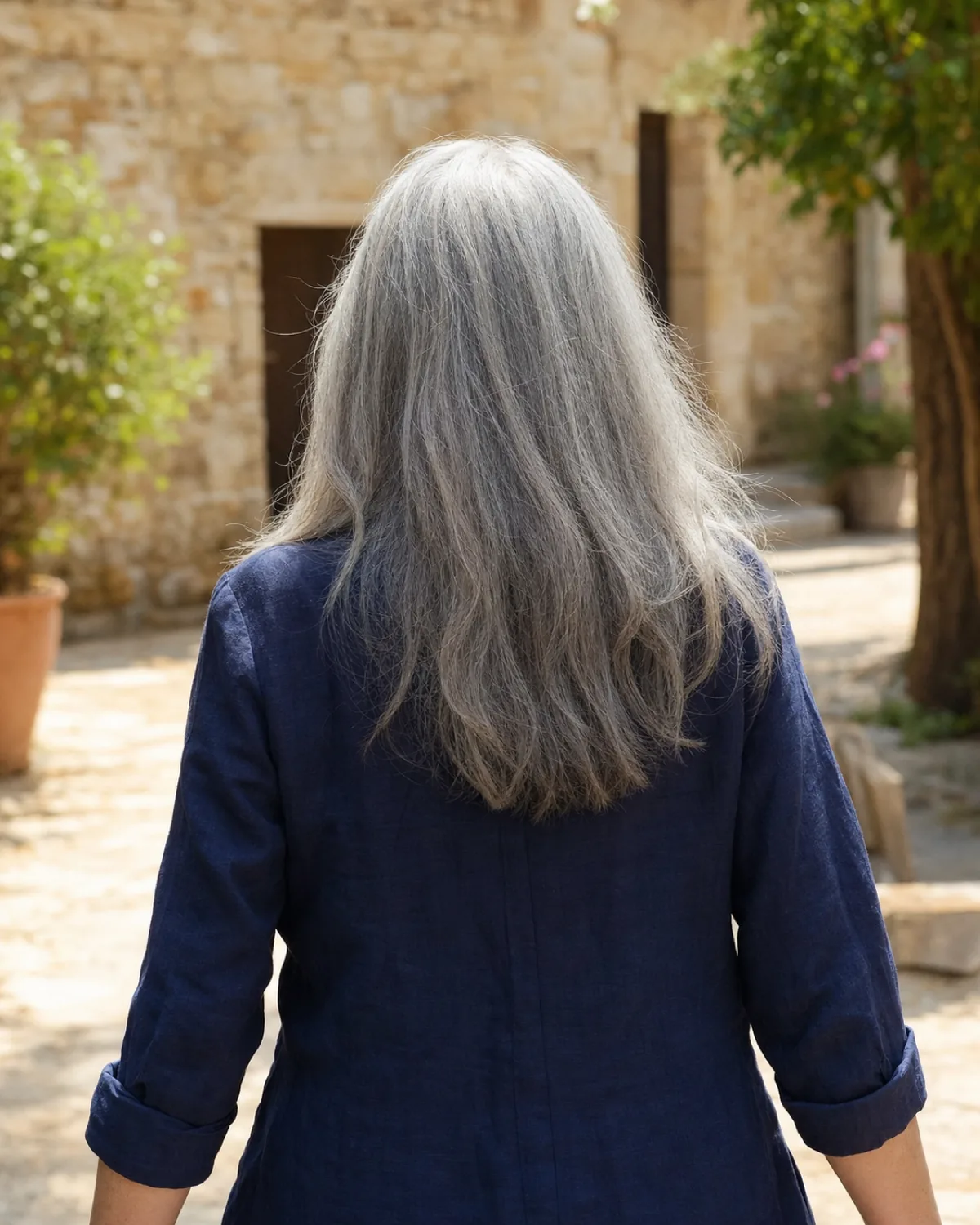 A woman walking away from the camera with long silver-grey hair falling past her shoulders to mid-back, thick and dense with a slight sway, wearing a navy linen jacket in a bright outdoor courtyard