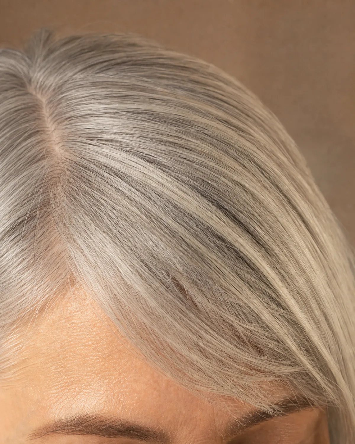 A close crop on the fringe and brow line of a woman with silver-grey hair, a side-swept fringe falling from a deep left-side parting, individual strands catching warm daylight against a soft taupe background