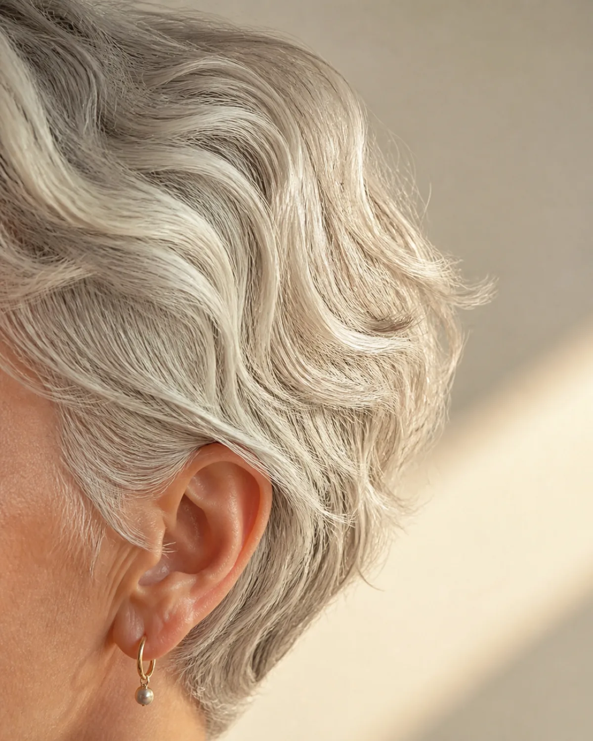 Close crop on the side of the head showing fine silver-white hair in a wavy pixie with visible S-curves occupying more space than straight hair would, golden window light against a cream-coloured wall