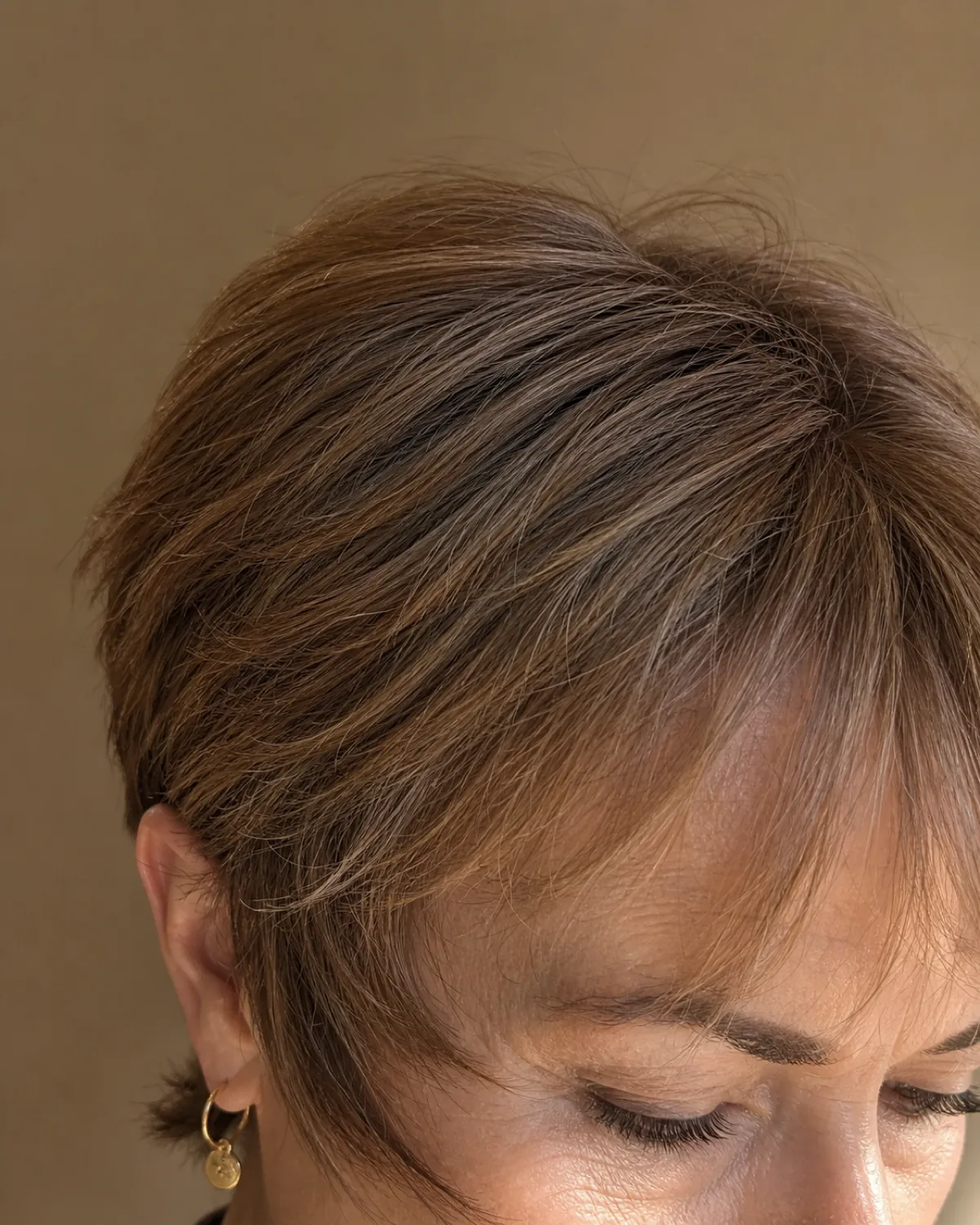 Close crop on the temple and brow area of a woman in her late 50s with fine honey-brown hair in a pixie, showing wispy feathered pieces extending in front of and below the ear, warm directional light against a sand-coloured wall