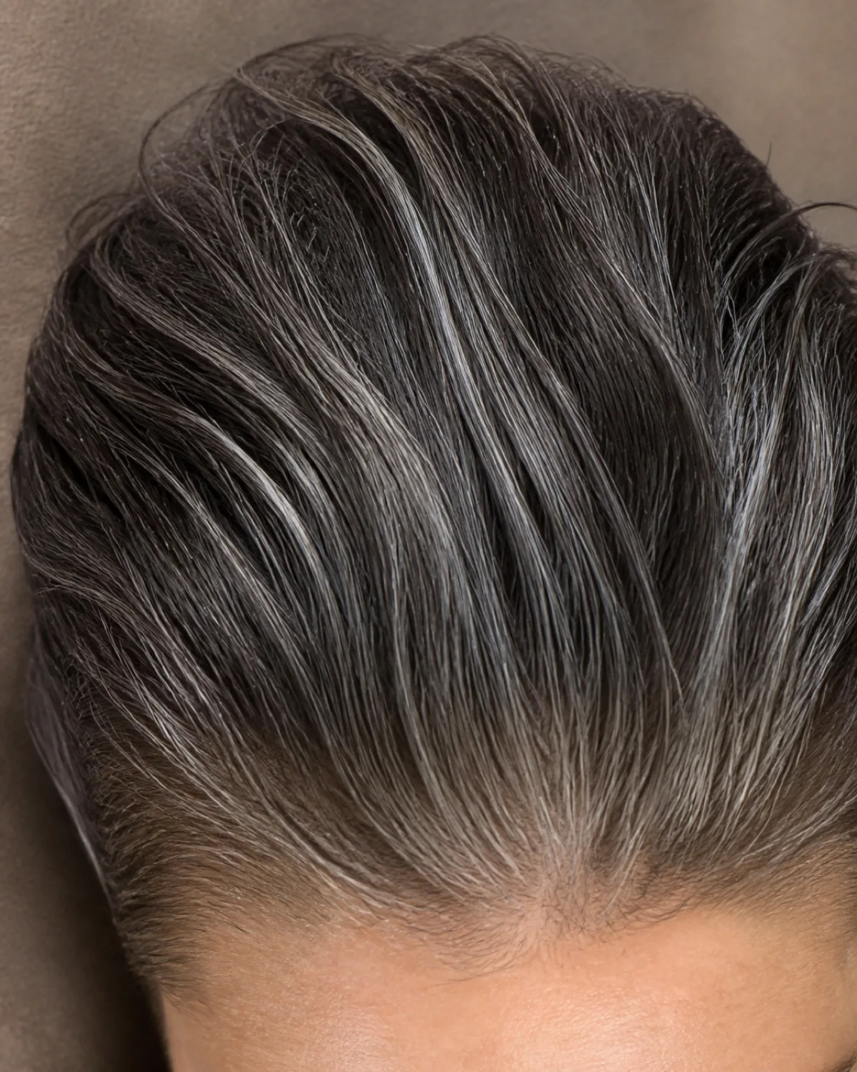 Top-down macro view of the part line and root area showing fine salt-and-pepper hair combed straight back in a slicked-back pixie with slight product sheen, individual strands visible at the roots against a warm taupe surface