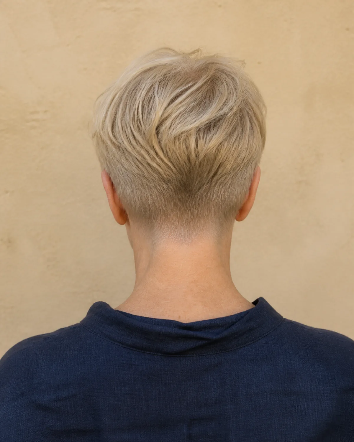 Shot from directly behind a woman in her late 50s with fine cool-blonde hair in a tapered nape pixie, showing the clean gradual taper from crown length down to a close-cropped neckline, overcast daylight against a warm linen-coloured wall
