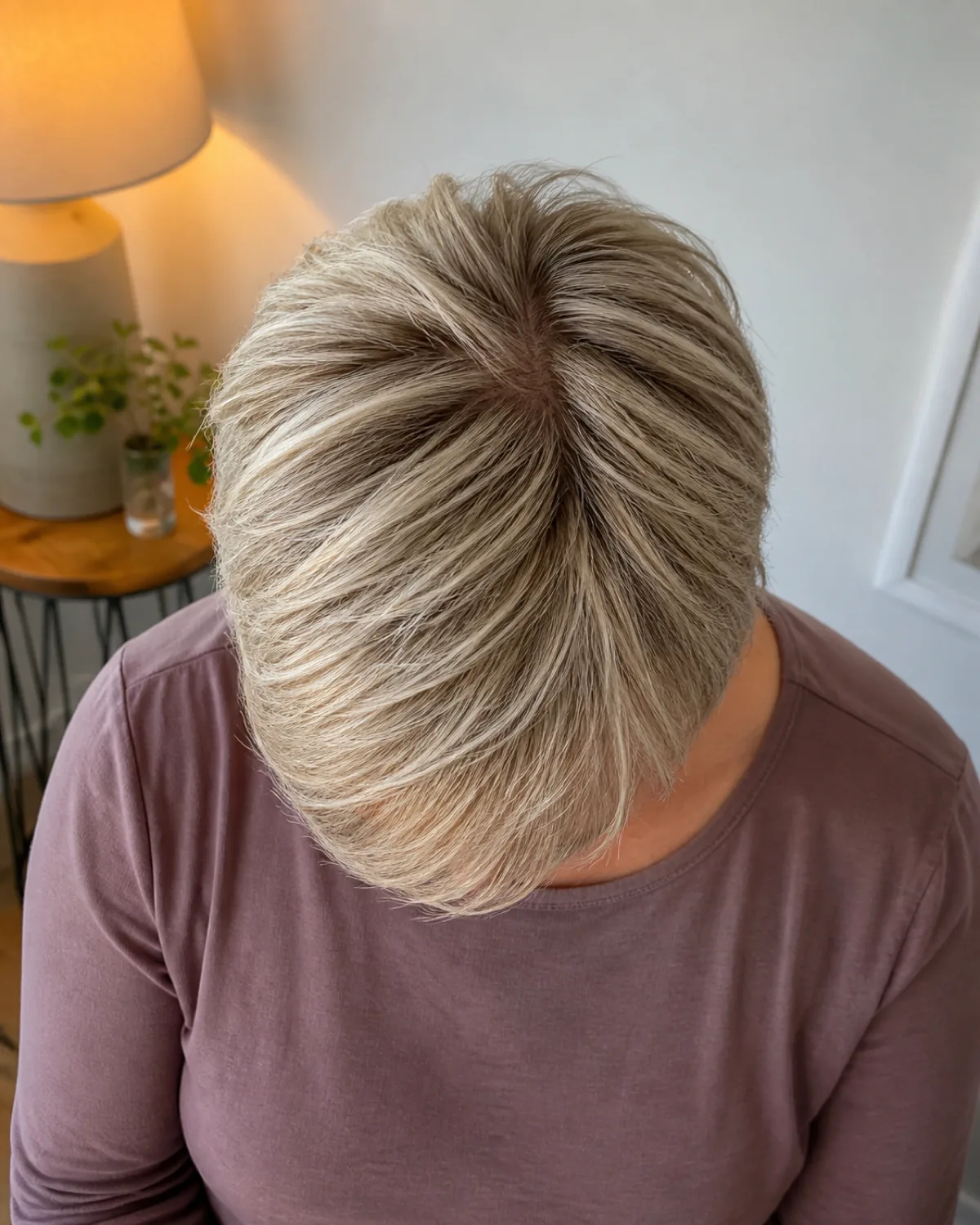Top-down view of a woman's crown showing fine ash-blonde hair in a textured crop with micro-layers lifting slightly from the scalp, warm lamp light against a soft white wall