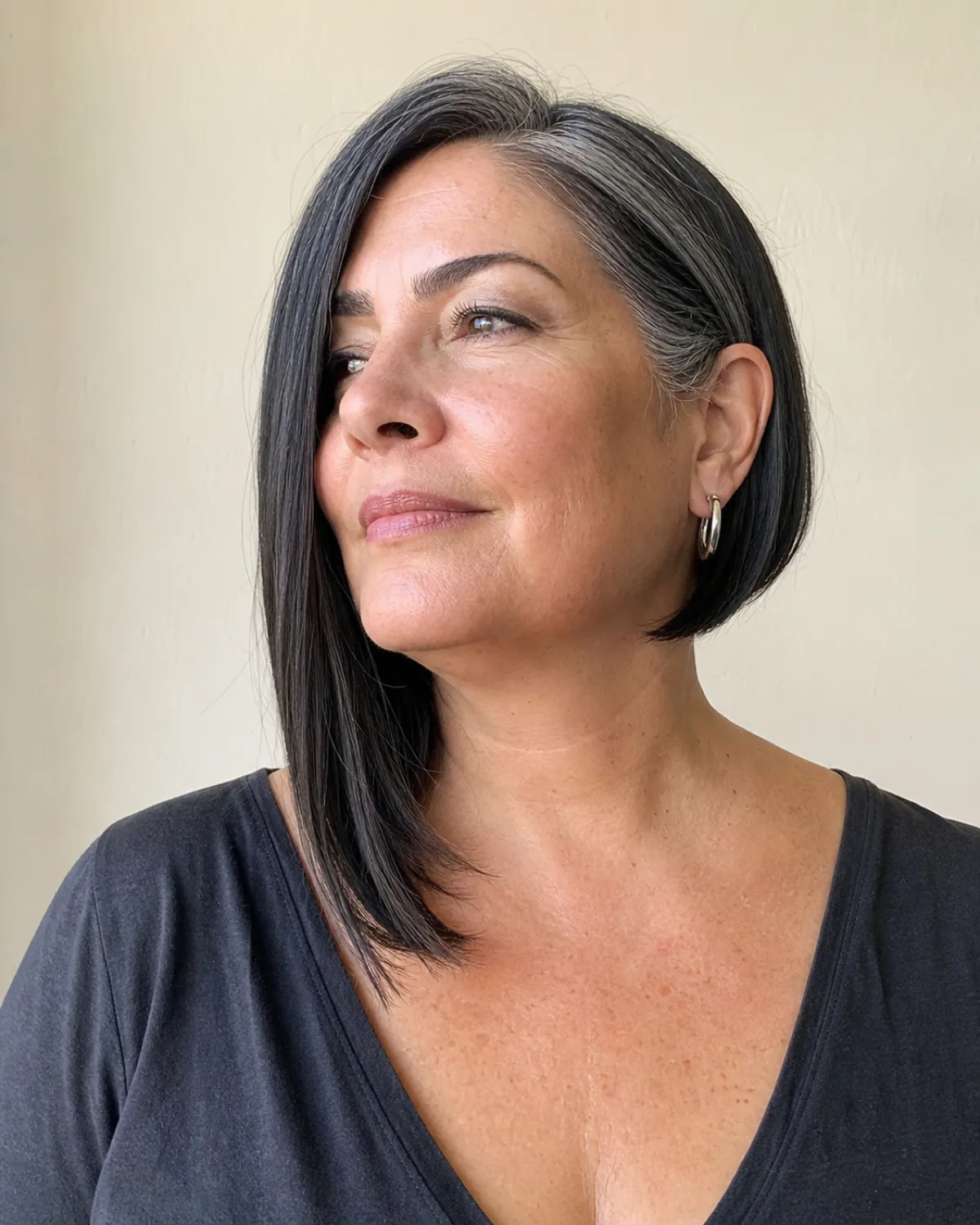 A woman over 50 with a fuller build in three-quarter turn to the left with chin up, dark hair with silver streaks in an asymmetric long bob with the camera-facing side noticeably longer at collarbone length and the far side shorter at the jaw, charcoal V-neck top, cream wall