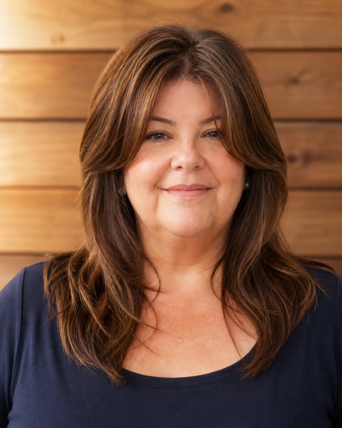 A woman over 50 with a fuller build and a wider jaw, front-facing with soft focus features, chestnut-brown hair with shorter face-framing pieces at the cheekbones and longer main length past the shoulders, navy scoop-neck top, wood-panelled wall