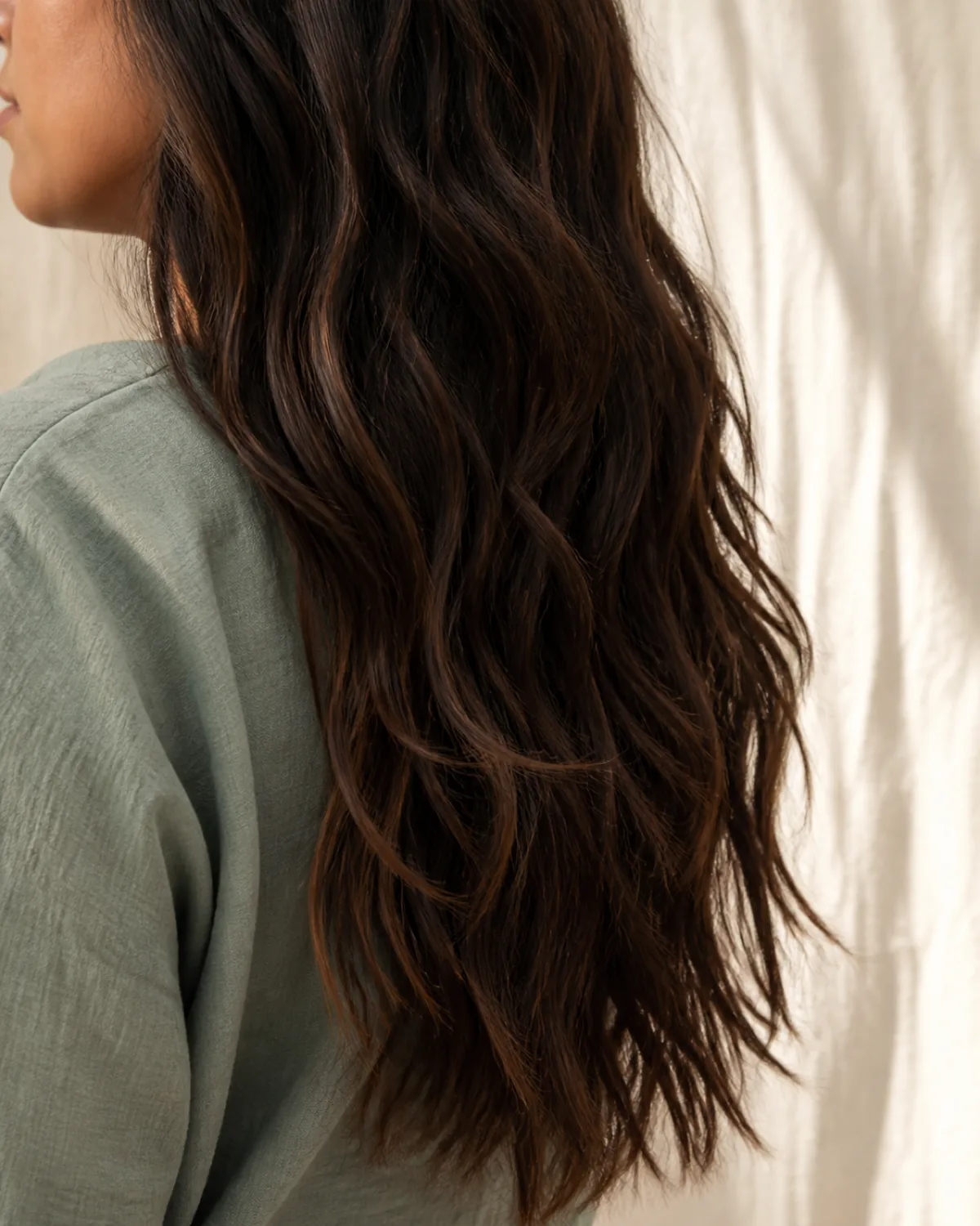 Close crop of long dark brown hair with warm highlights falling past the shoulders in loose natural waves, individual strands visible at the ends with good density, shoulder in sage top partially visible, warm daylight, linen background